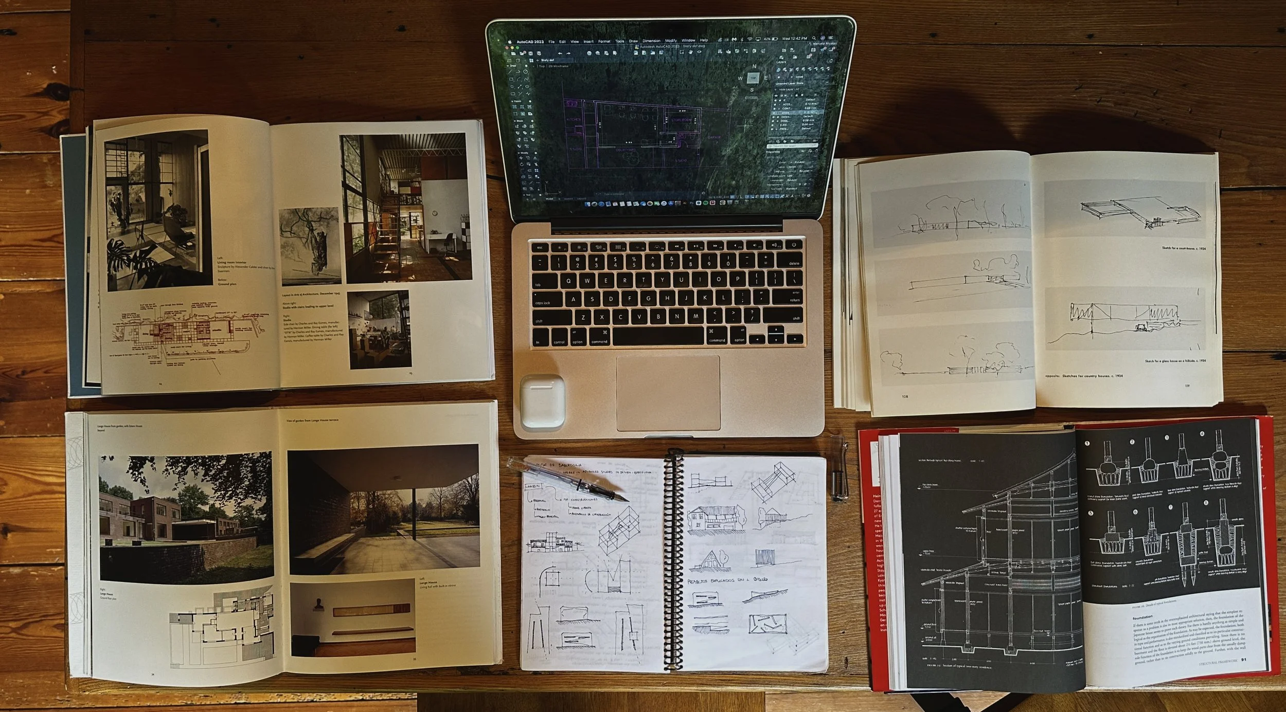 A top-down view of a wooden desk with open books, notebooks, a laptop displaying architectural design software, pens, and sketchpads, all related to architecture and design.