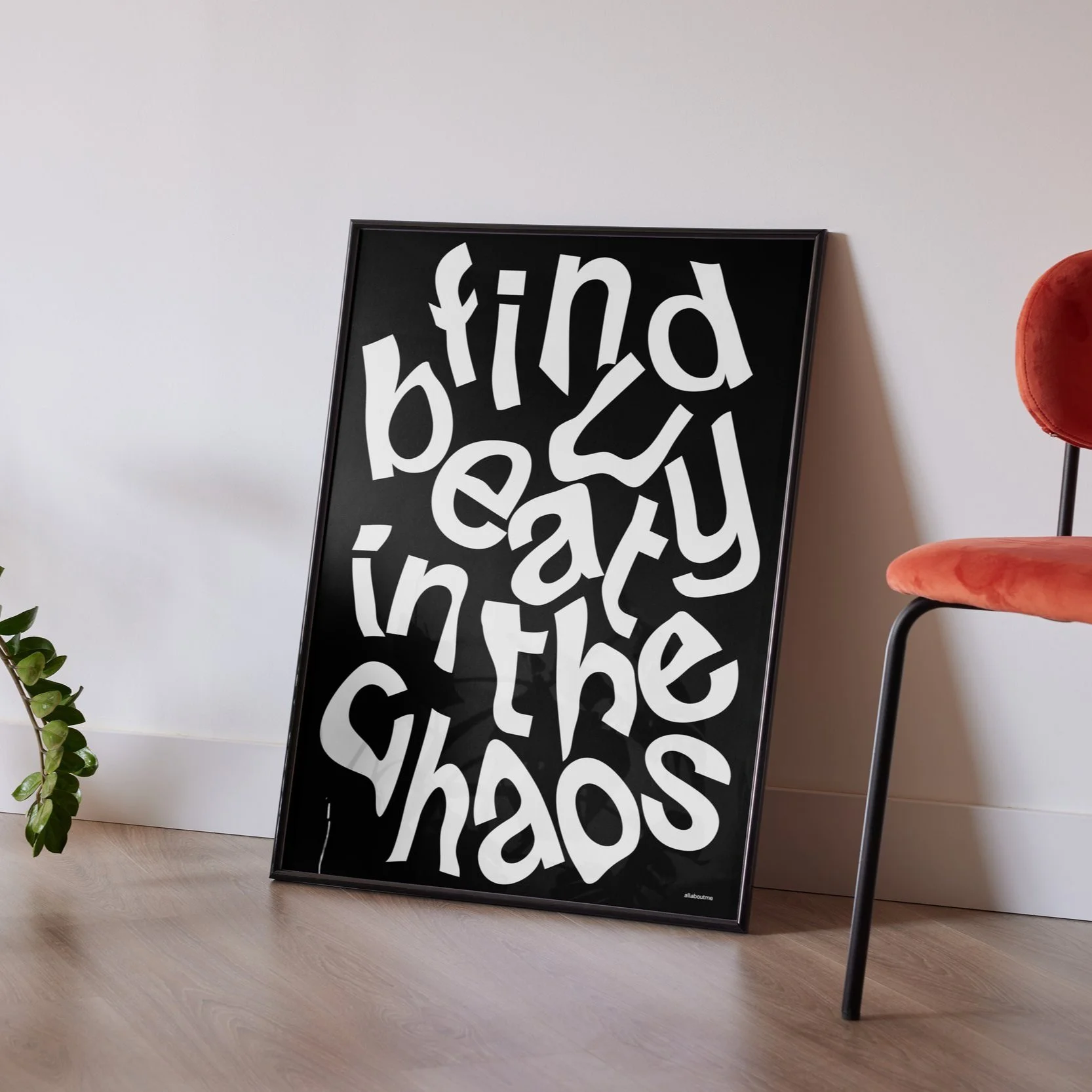 Framed black poster with white, distorted lettering that reads "find beauty in the chaos," leaning against a plain white wall on a light wood floor. A red chair is partially visible to the right, and a small green plant is on the left.