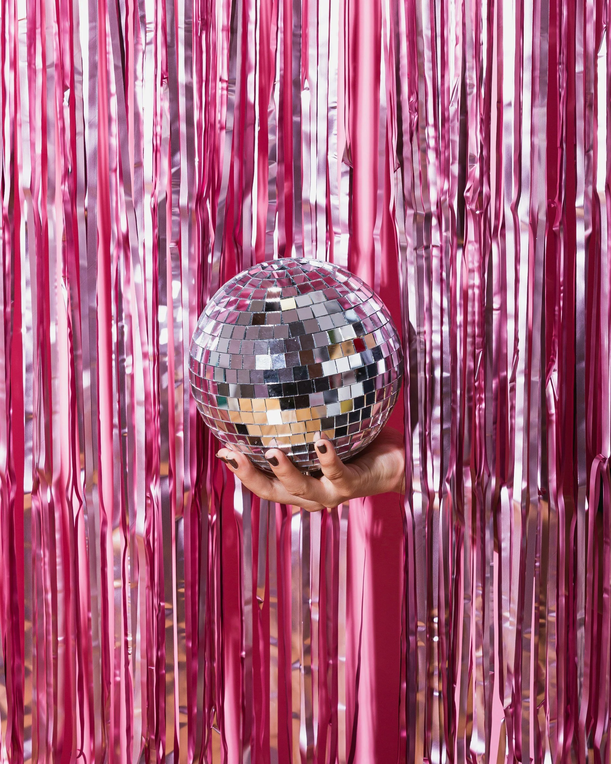 Person holding a silver disco ball in front of pink metallic fringe curtains.