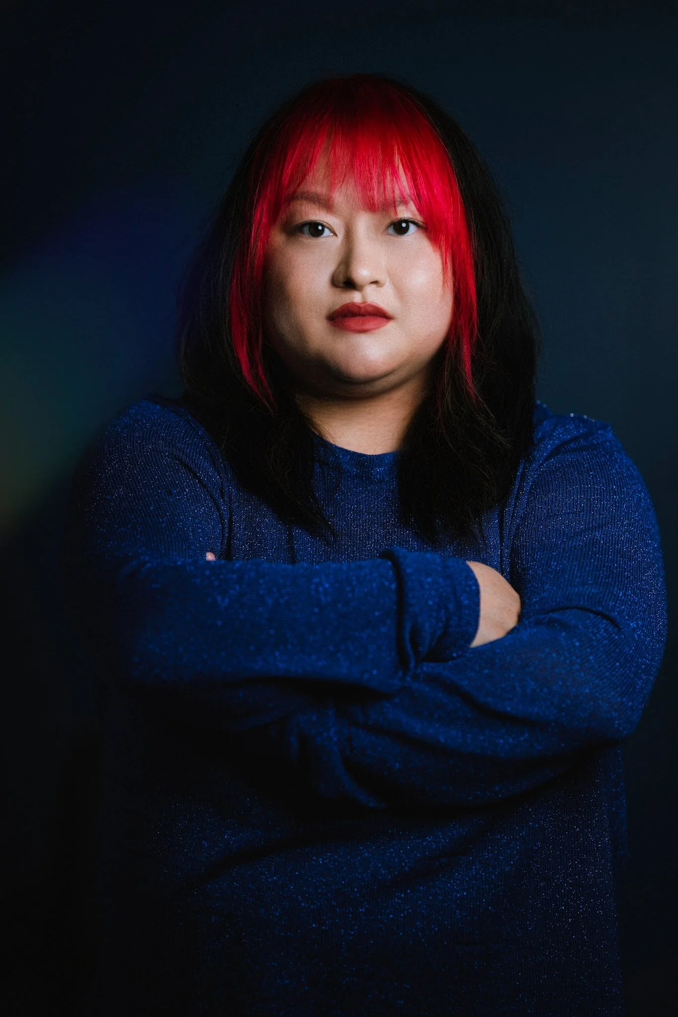 Portrait of a woman with black and red hair, wearing a sparkly blue top, crossing her arms, against a dark background.
