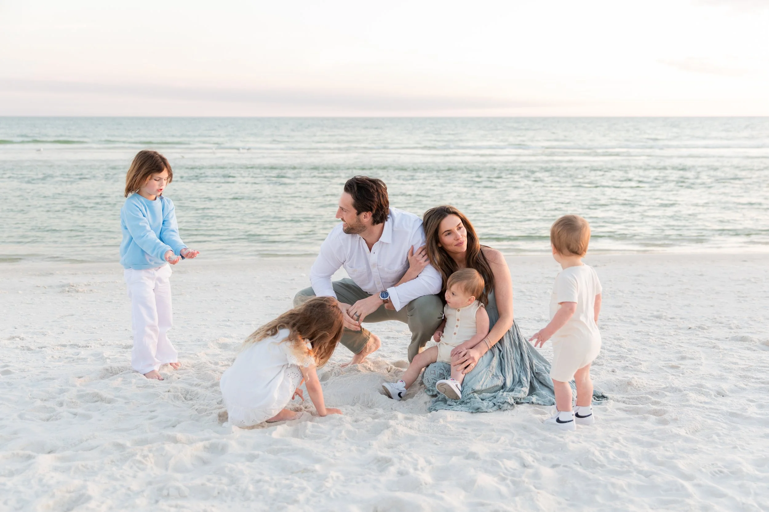 A family enjoys a day at the beach, with two adults and four children, some playing in the sand and others standing or sitting on the white sandy beach by the ocean during early evening or sunset.