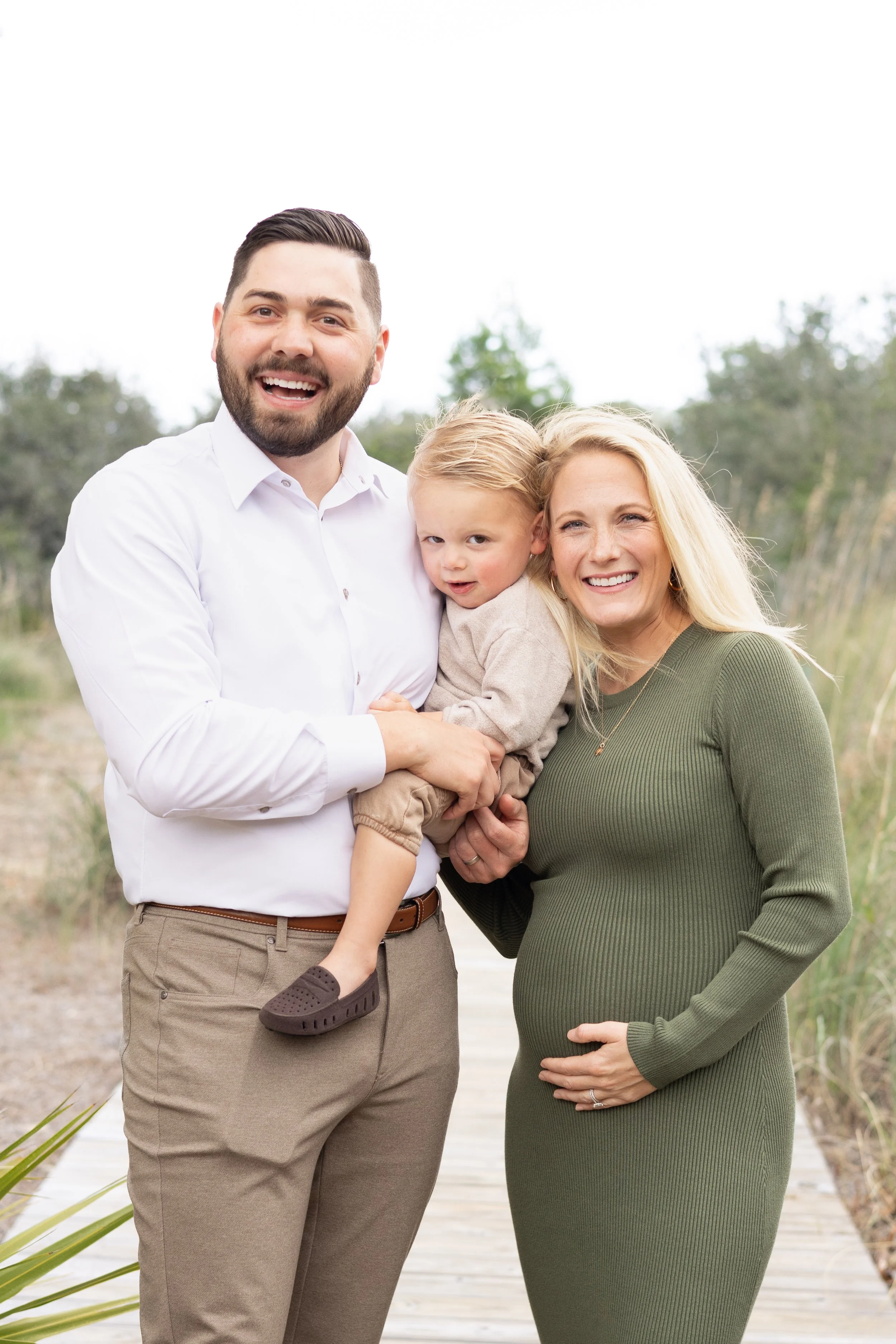 Happy family of three outdoors on a walkway, with a man holding a young child and a woman pregnant, all smiling.