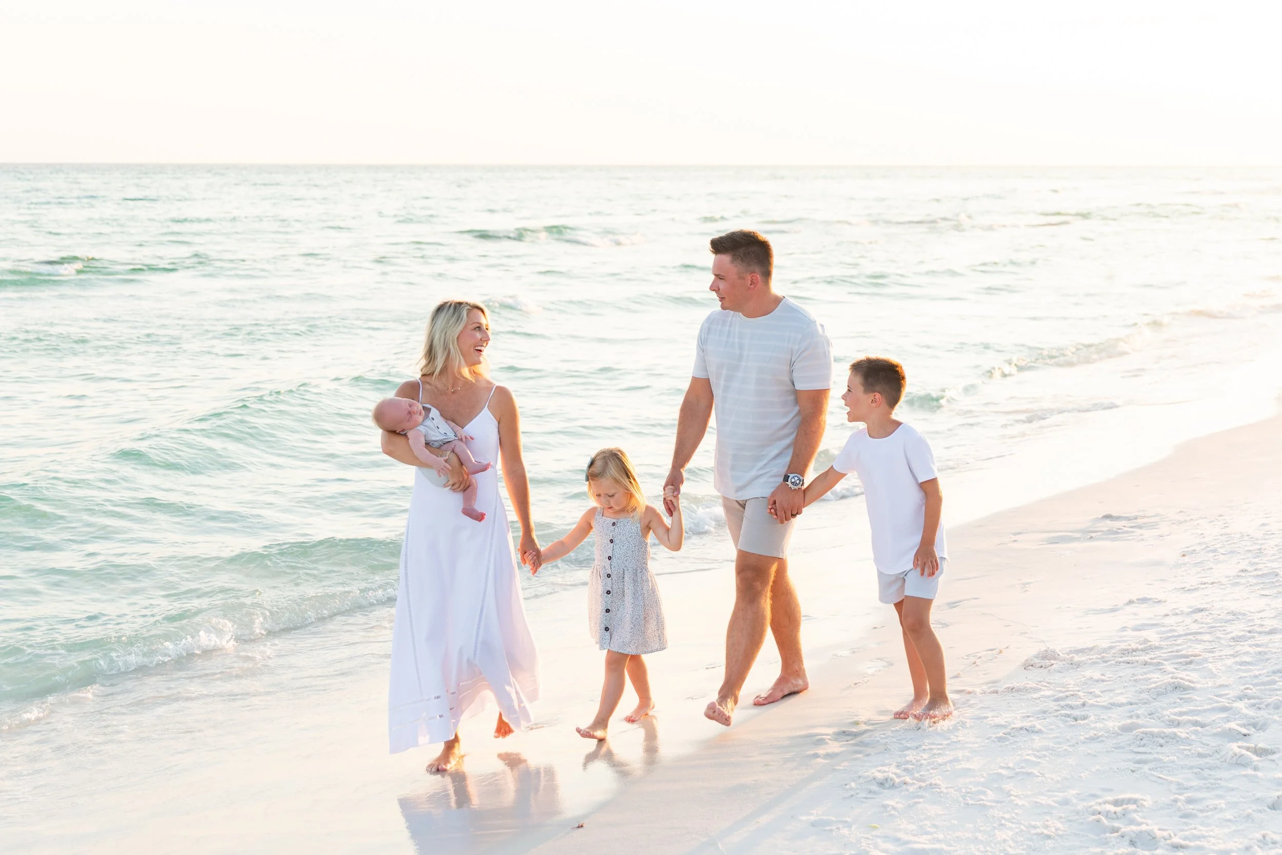A family of five holding hands and walking on the beach during sunset, with ocean waves in the background.
