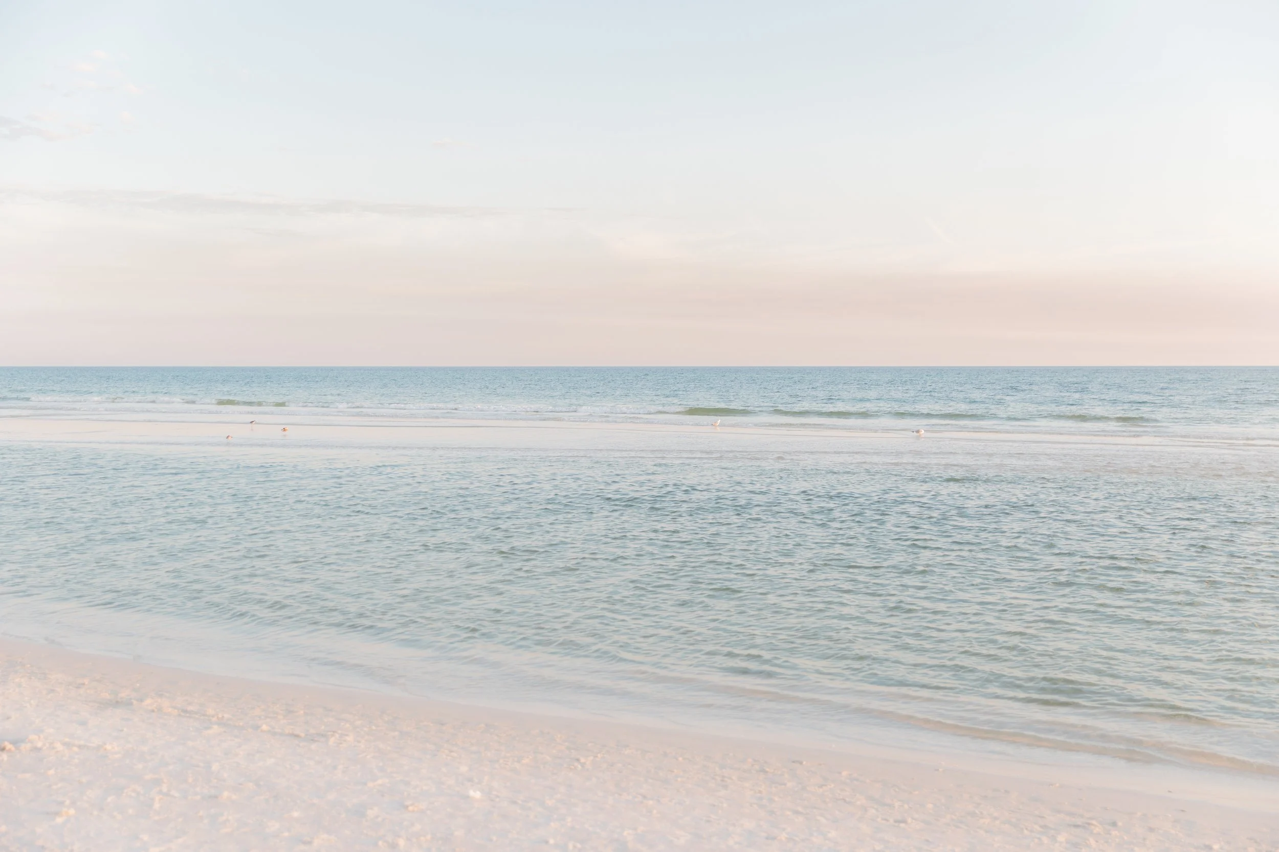 Calm ocean waves gently lapping onto a sandy beach under a sky with soft clouds during sunrise or sunset.