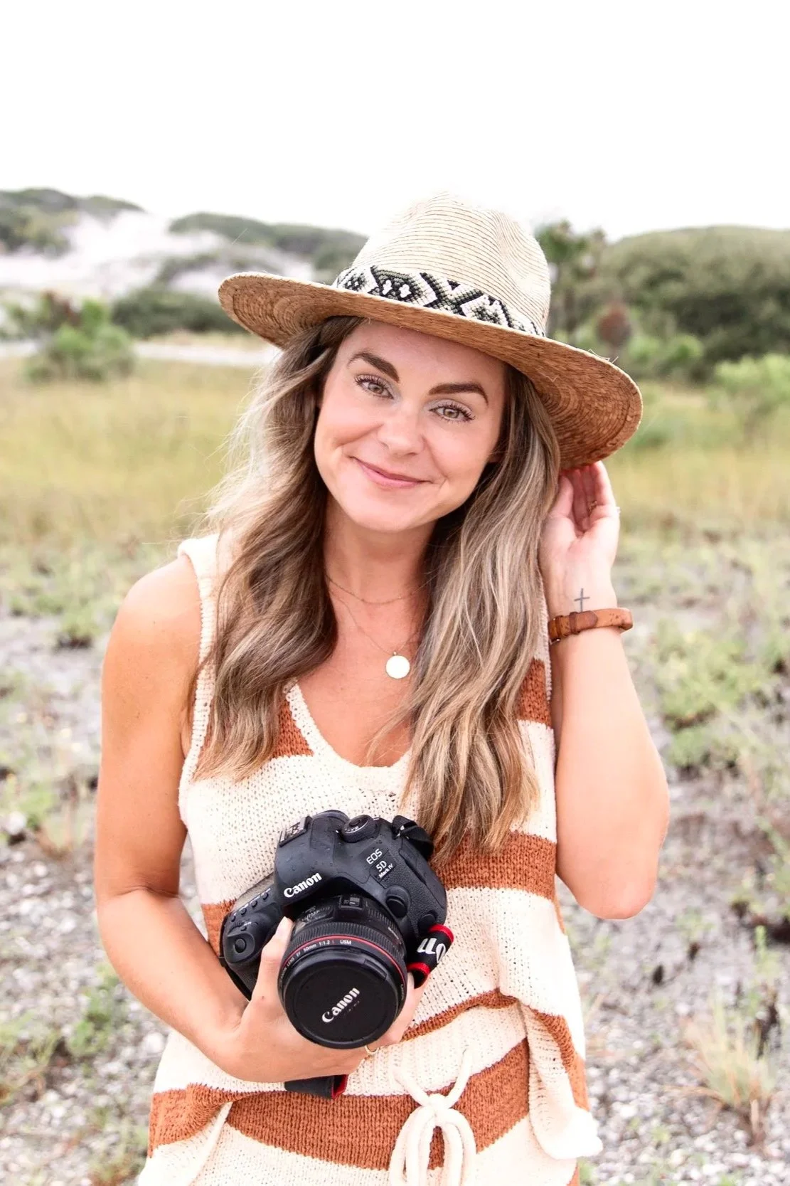 Young woman smiling, wearing a wide-brimmed straw hat and striped sleeveless dress, holding a Canon camera, standing outdoors in a grassy landscape.