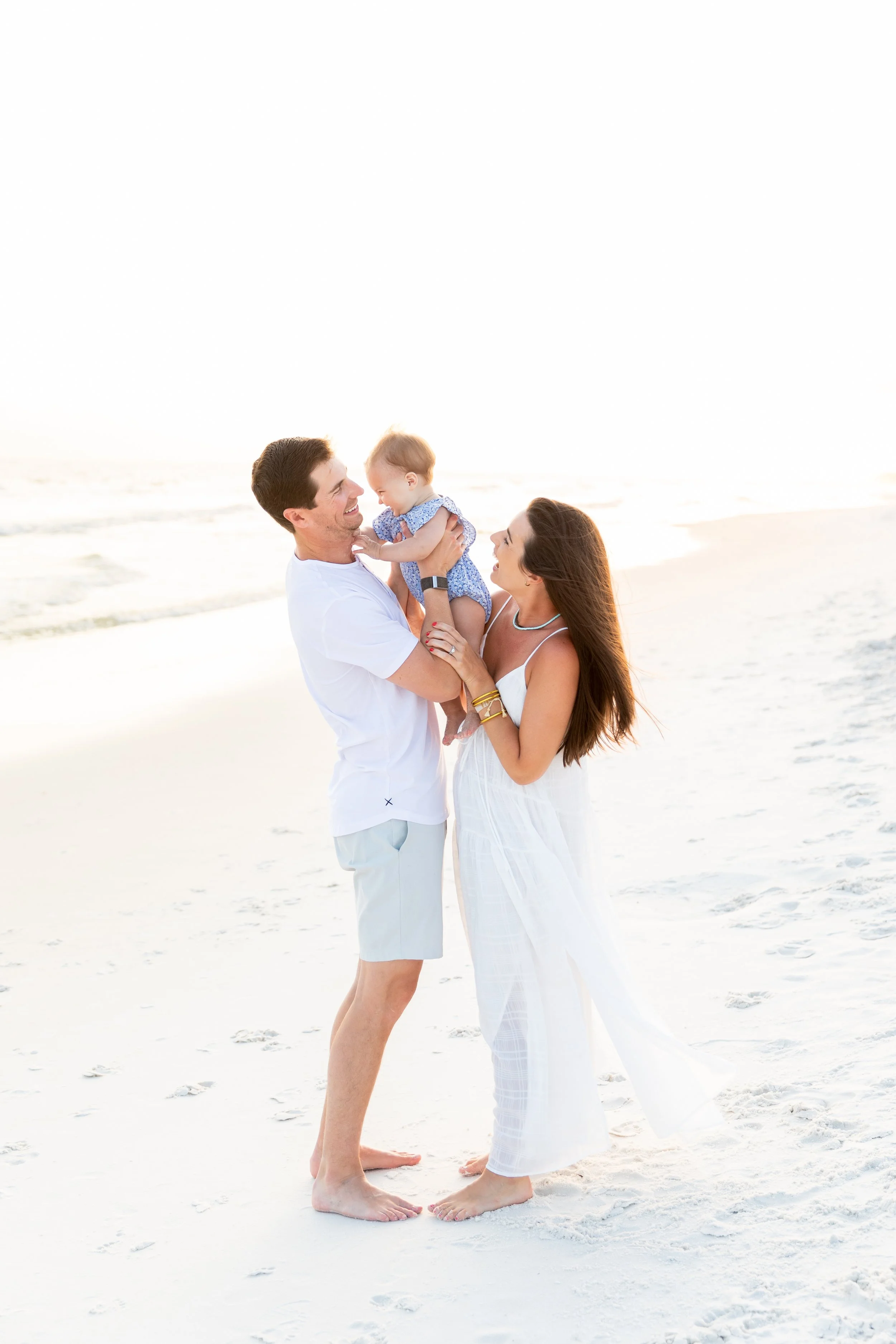 A family of three at the beach, with the father holding a baby girl while the mother interacts with them, all smiling in a bright, sunny setting.