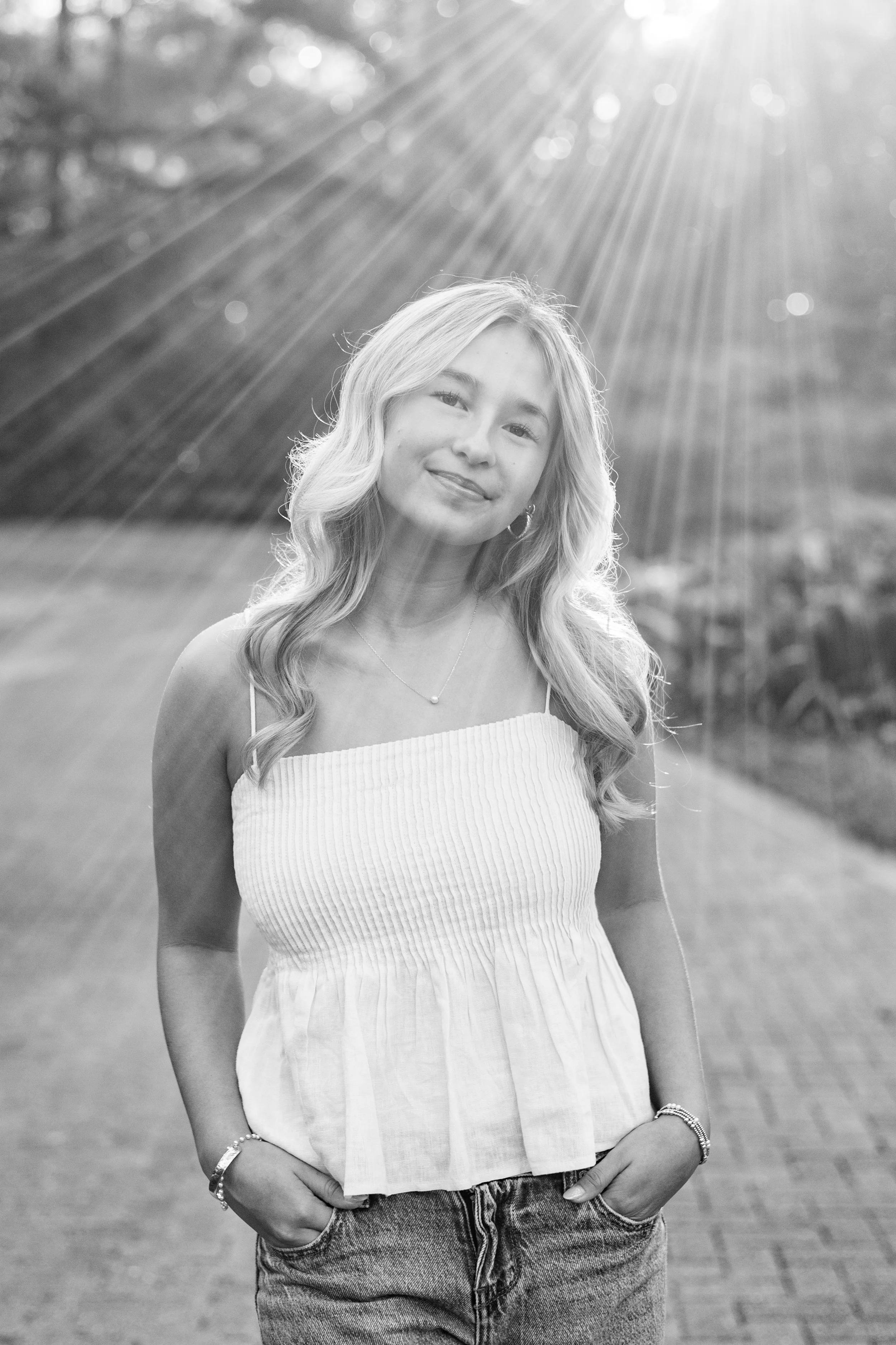 Black-and-white photo of a young woman smiling outdoors, with sunlight streaming down behind her, wearing a sleeveless top and jeans, standing on a brick pathway.