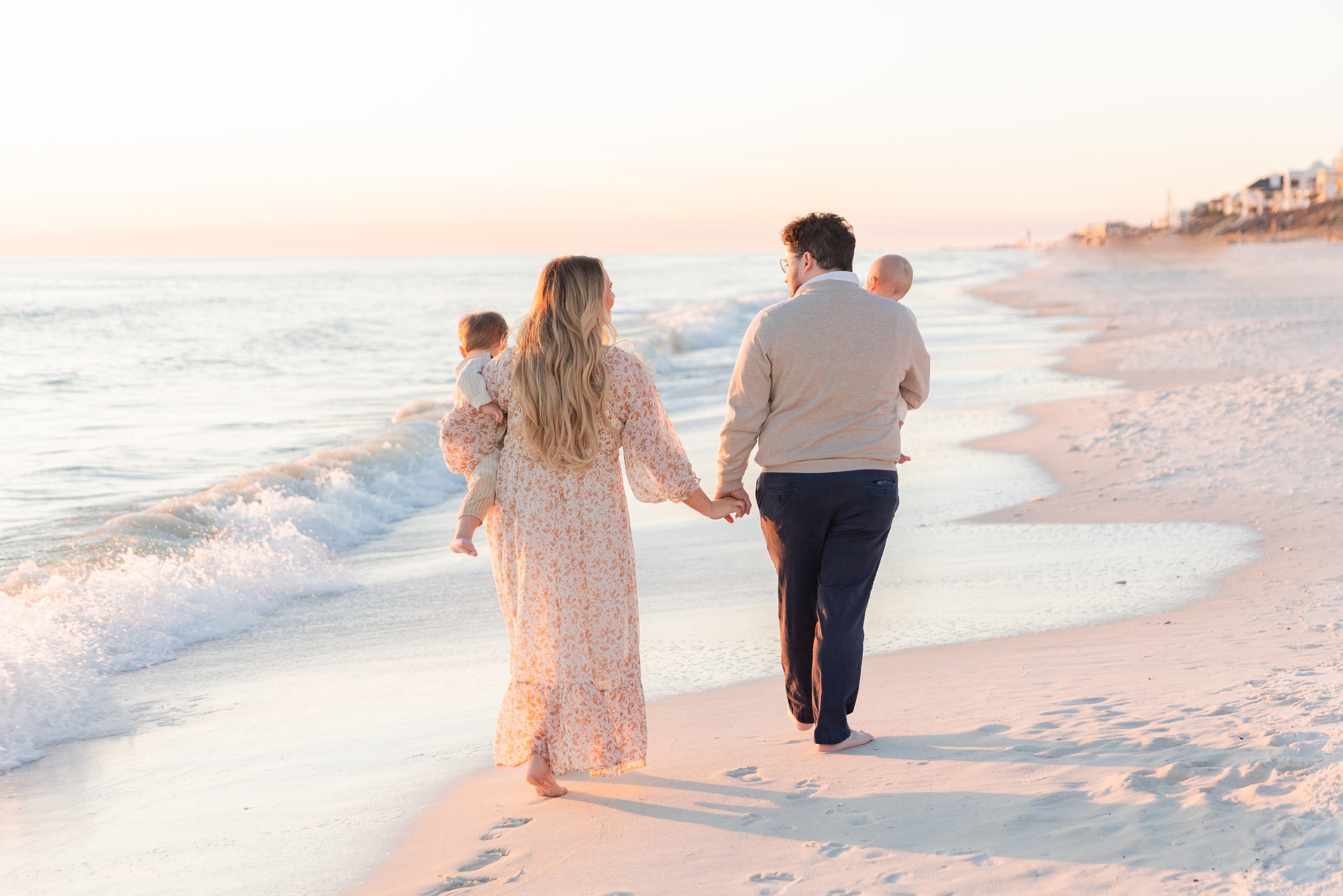 A happy family of four walking on a sandy beach during sunset. The mother is holding hands with the father, and they are carrying their two young children; one child is in the mother's arms and the other is in the father's arms. The beach has gentle 