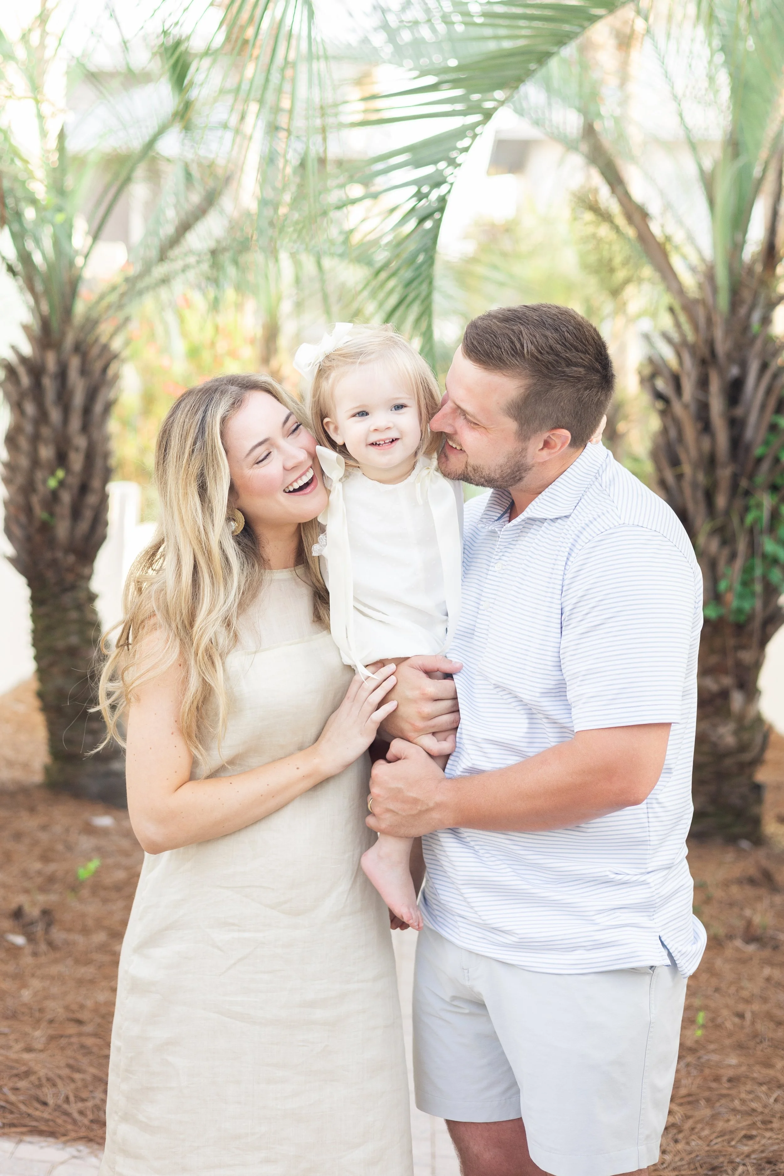 A happy family of three, a mother, father, and their young daughter, enjoying time outdoors among palm trees, smiling and laughing.