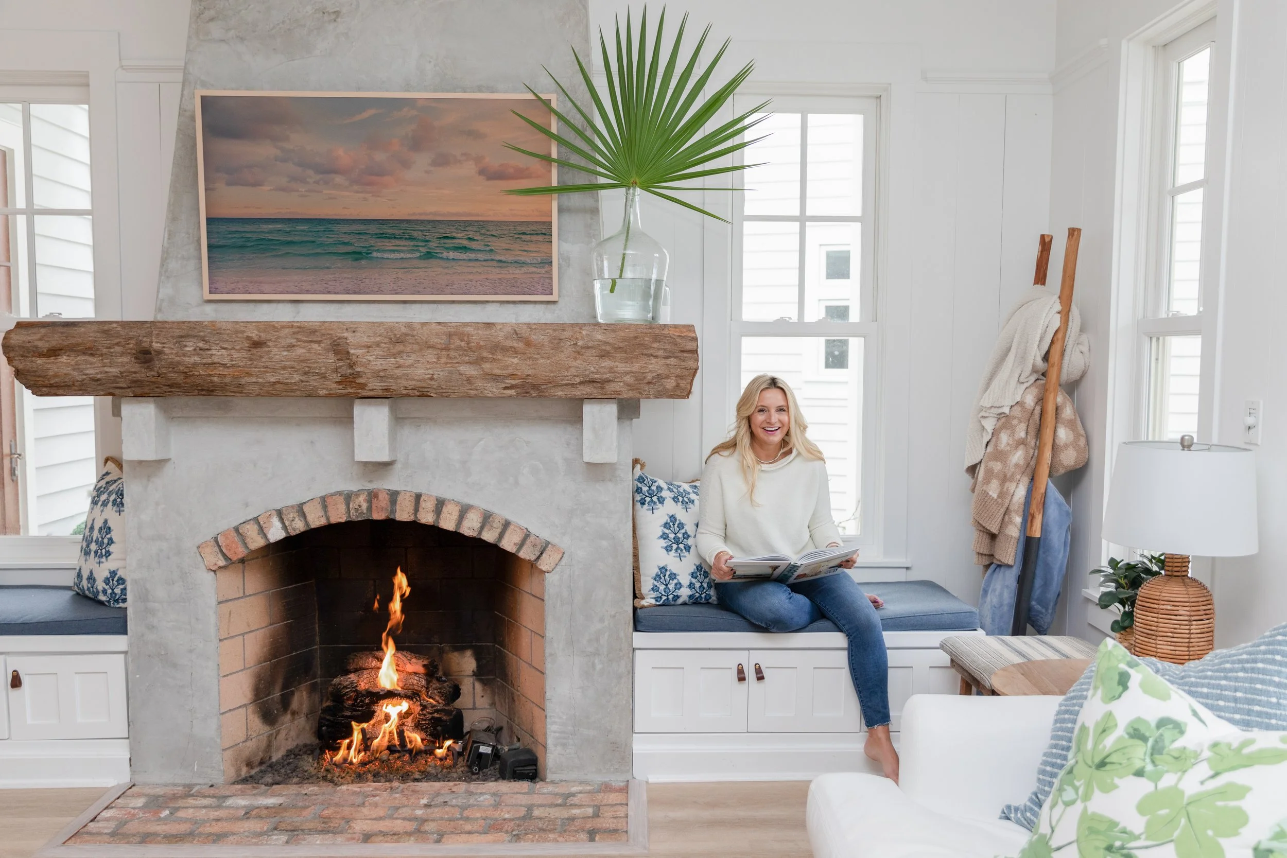 A woman sitting on a window seat near a fireplace with a fire, holding a magazine, in a bright, cozy living room with white walls, a large painting of the ocean, and decorative pillows.