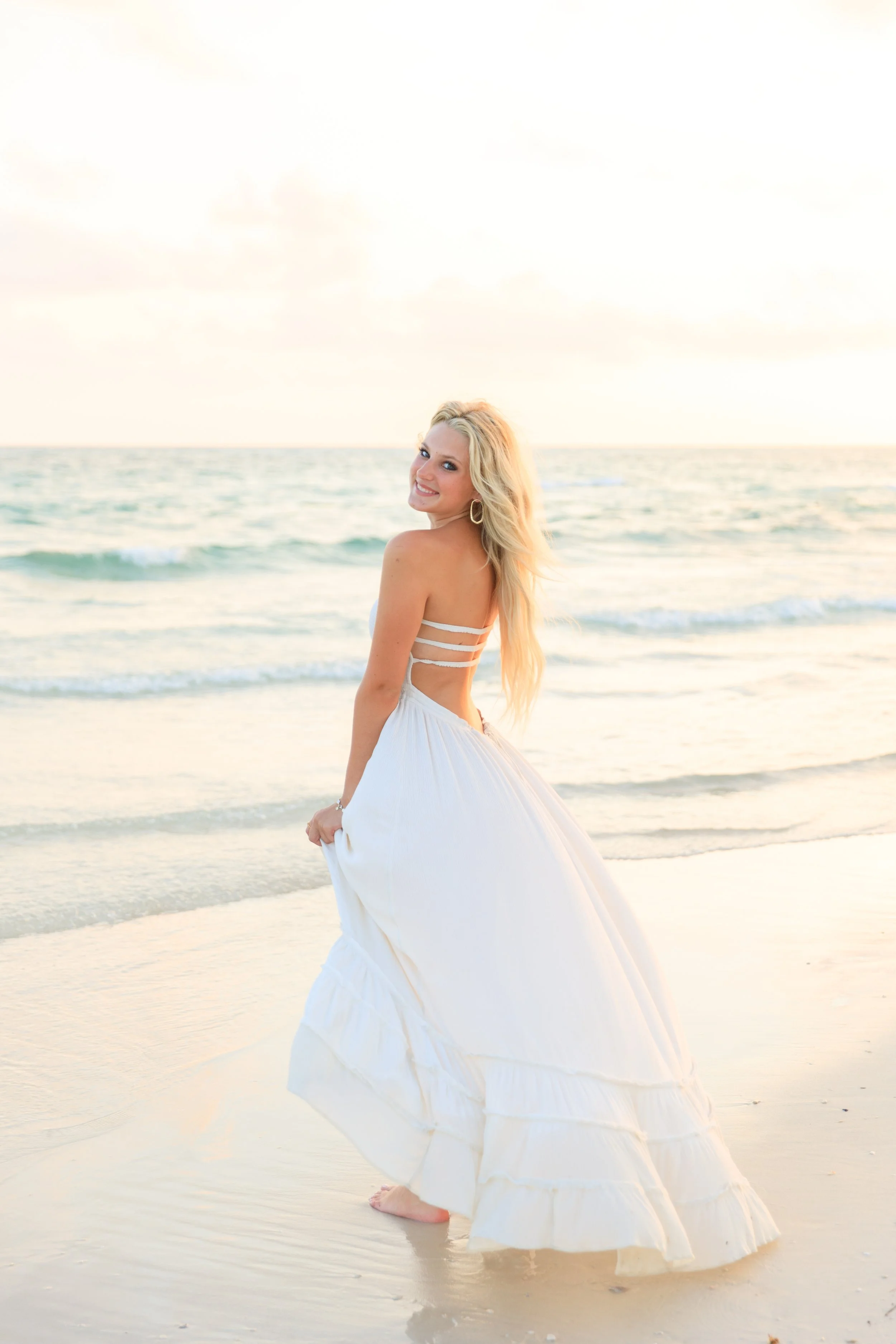 A woman with long blonde hair wearing a white dress with cutout details, standing in shallow water on a beach during sunset, smiling at the camera.