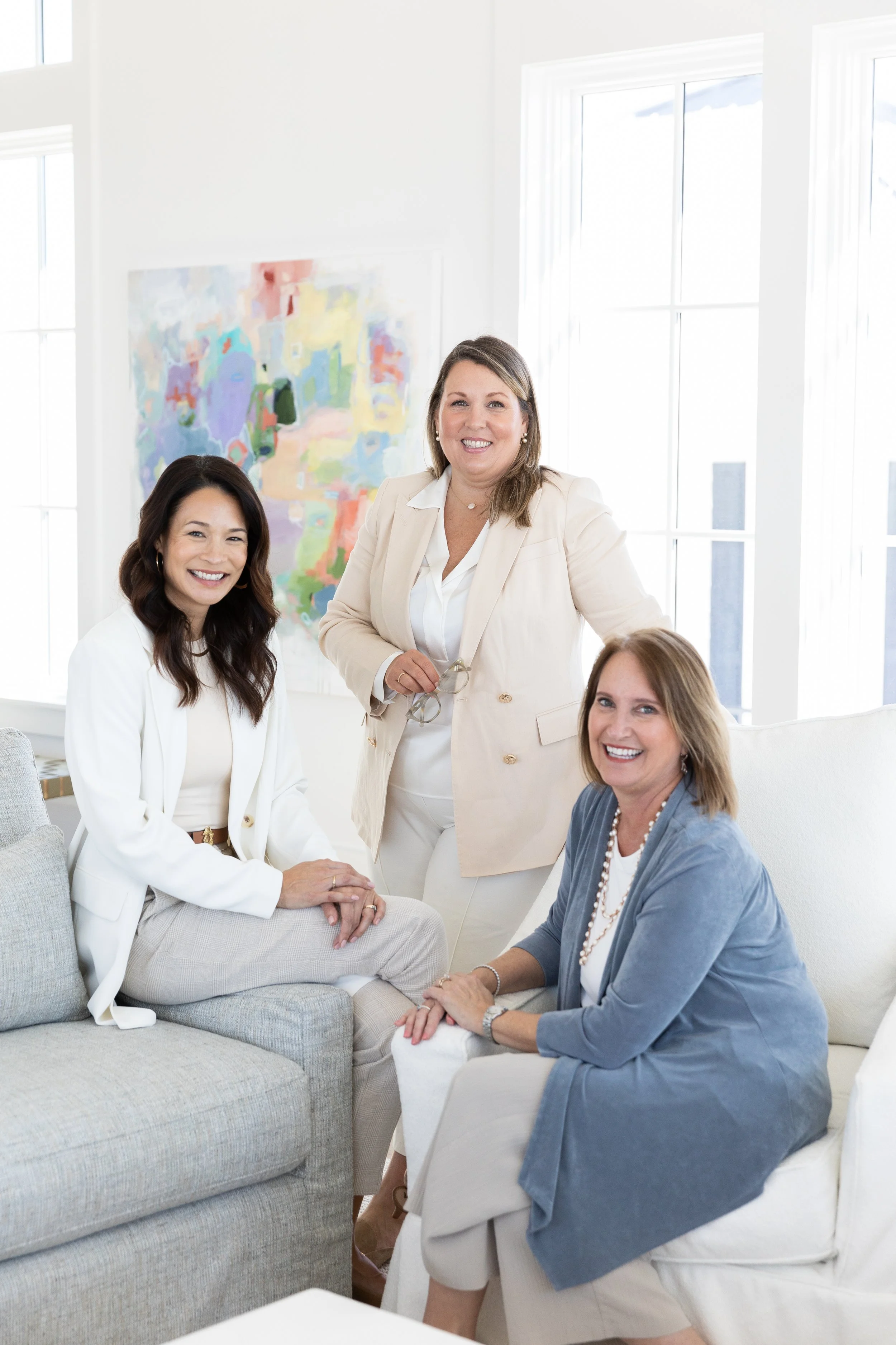 Three women in a bright, modern living room, smiling and posing for the camera, with large windows and an abstract painting in the background.
