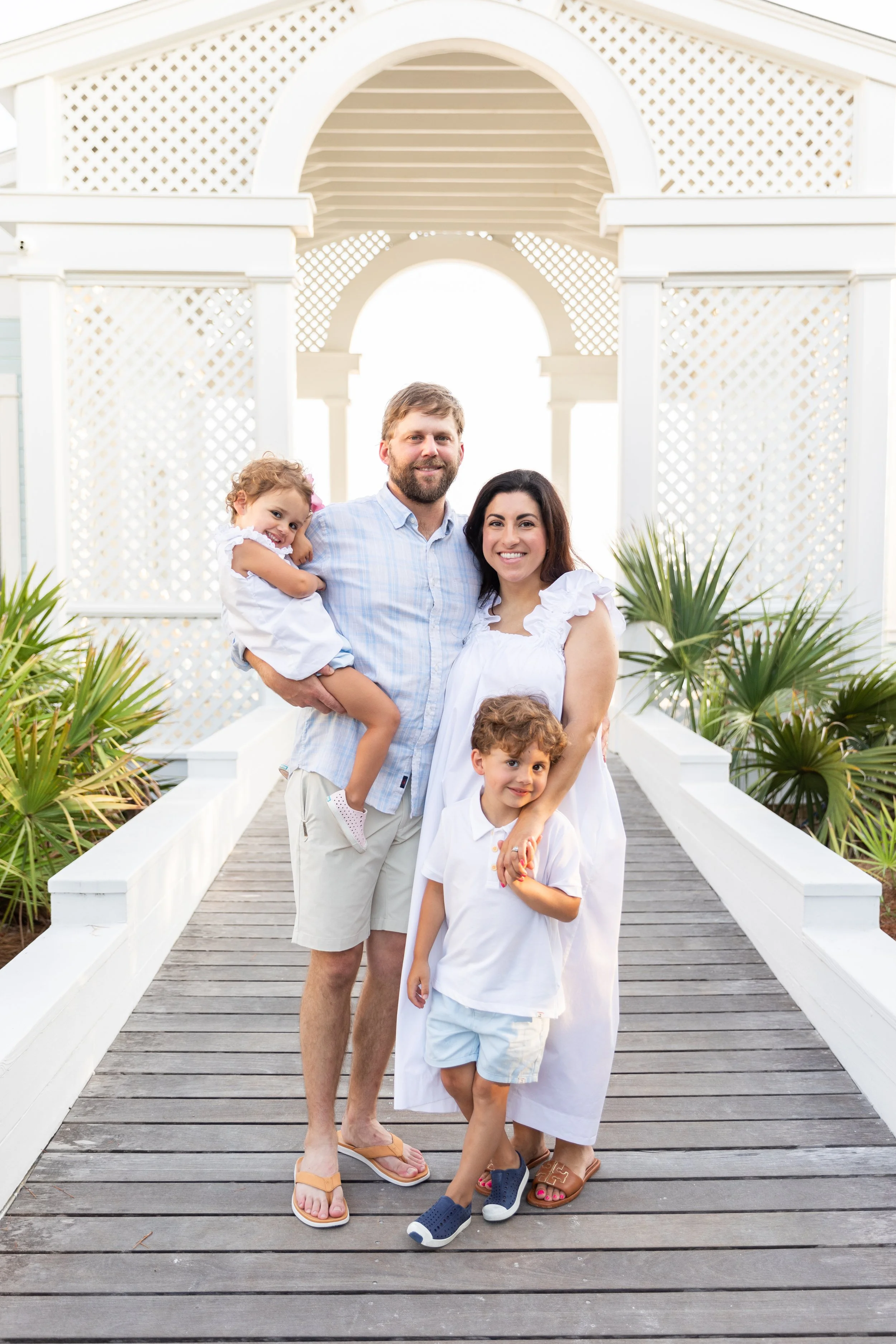 A happy family of four, includes a man, woman, and two children, standing on a wooden walkway with white lattice and lush green plants in the background, dressed in light summer clothing.