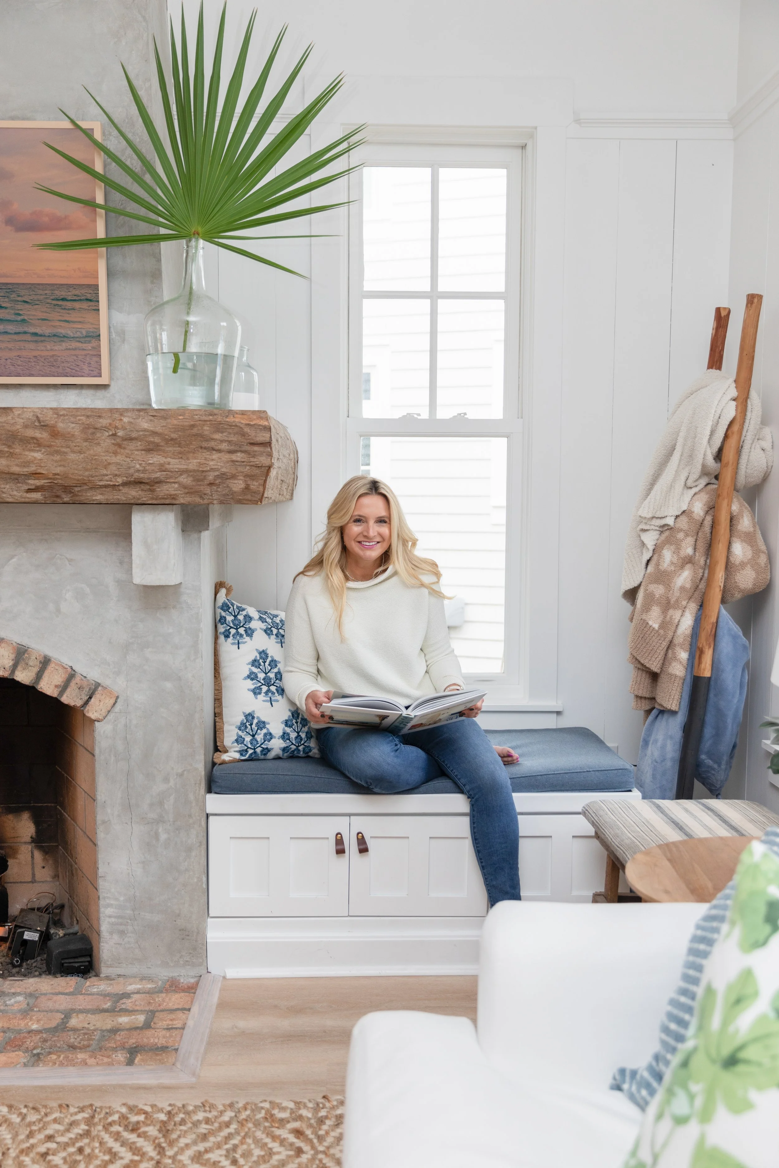 A woman sitting on a built-in window seat in a cozy living room, holding a book and smiling, with a large window, fireplace, and decorative elements around her.