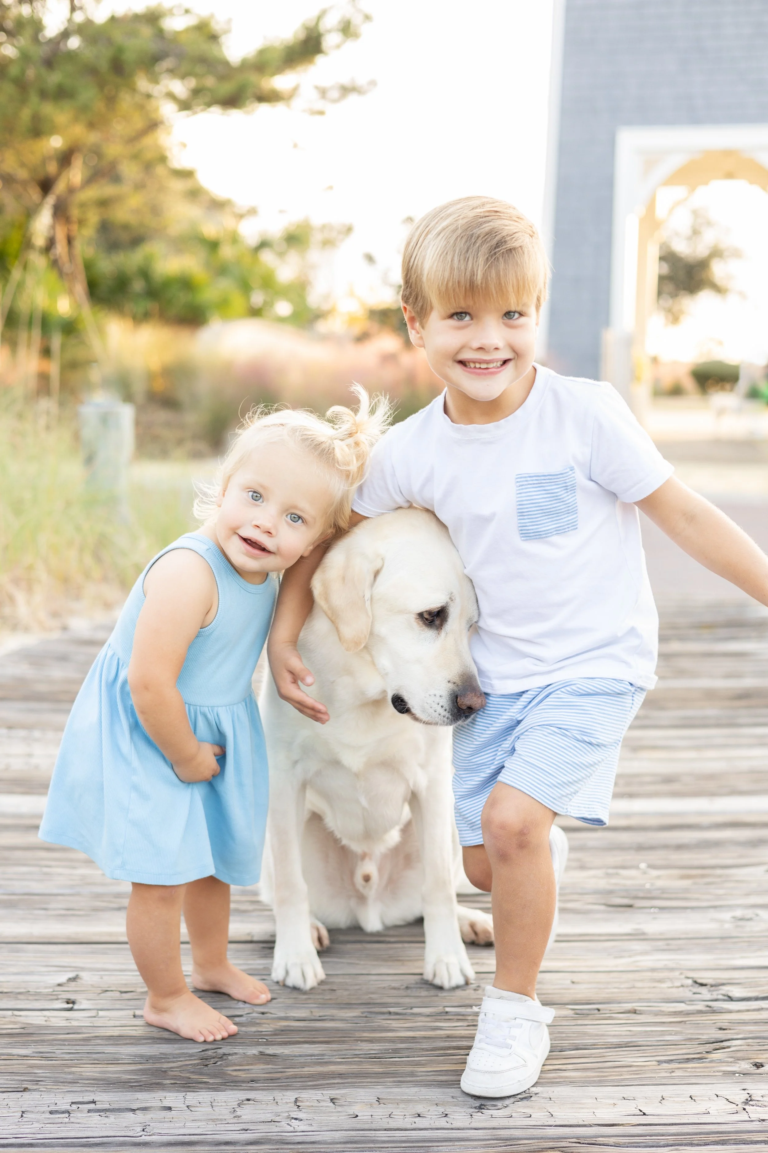 Two children, a boy and a girl, with a Labrador retriever puppy outdoors on a wooden pathway, smiling and posing for the camera, with trees and a building in the background.