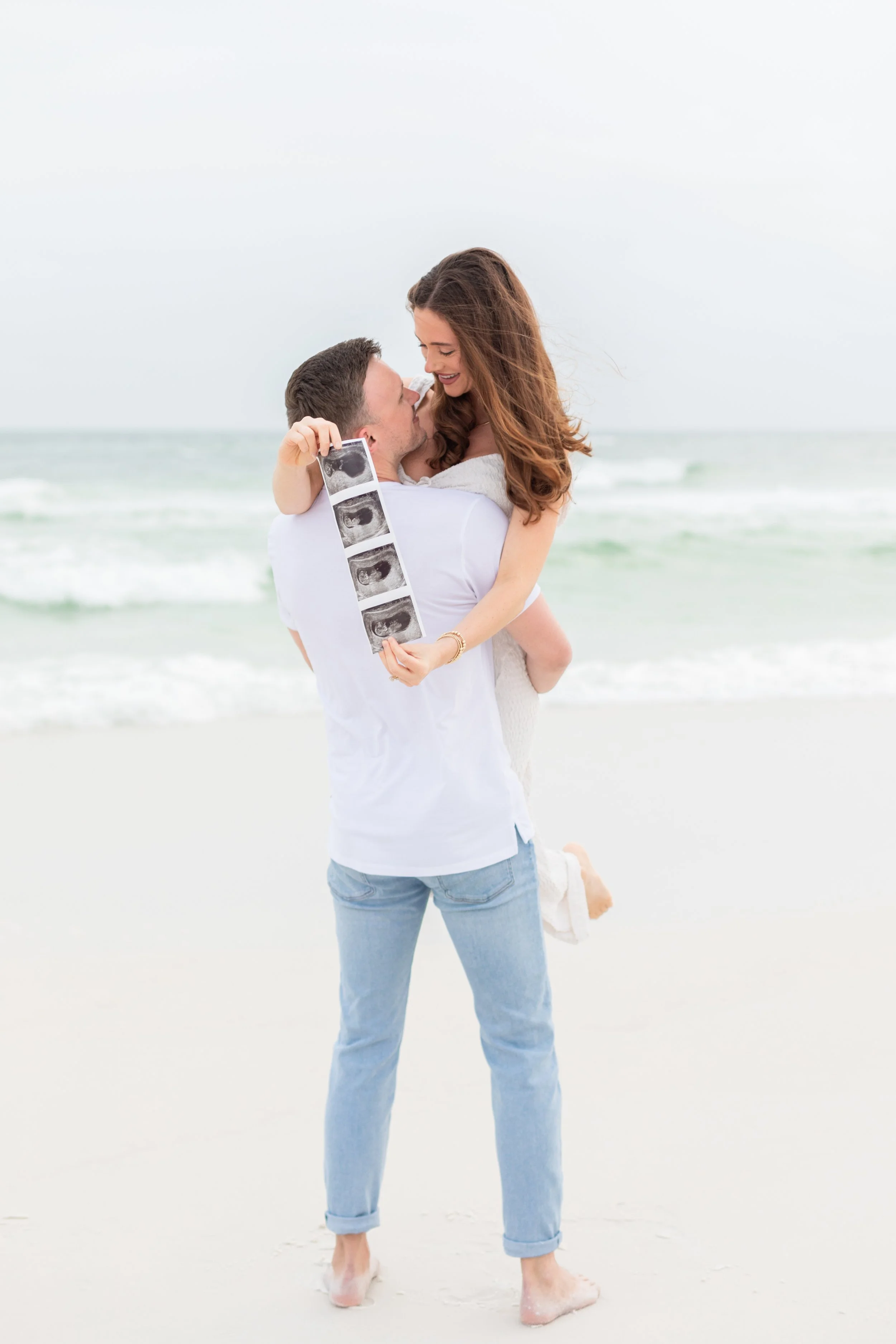 A couple on the beach holding an ultrasound photo of a baby, smiling and looking at each other.