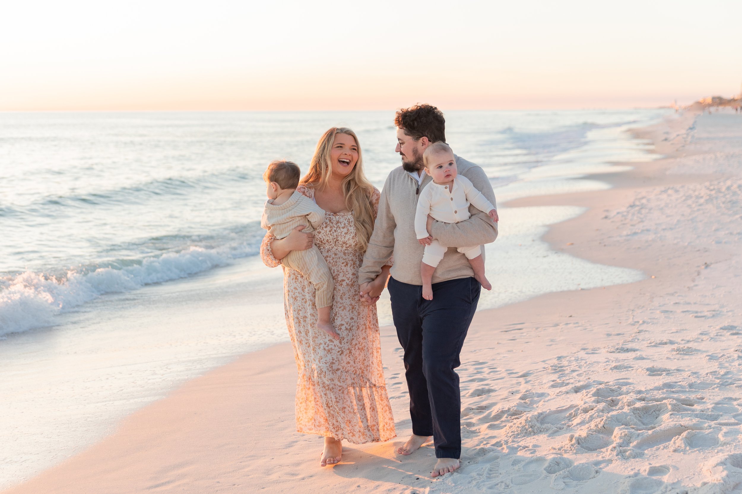 A family of four walking along the beach during sunset, holding hands and smiling.