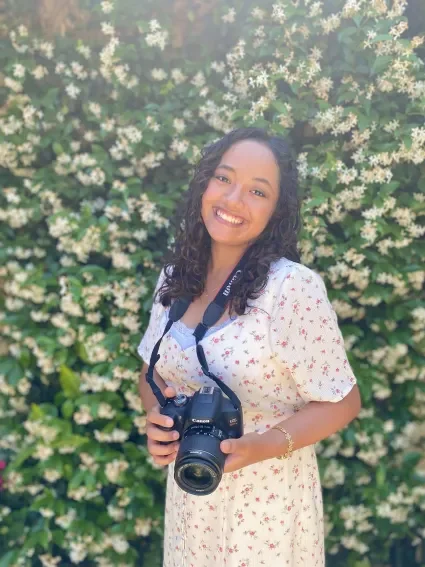 A smiling woman with curly hair holding a camera stands in front of a bush with white flowers.