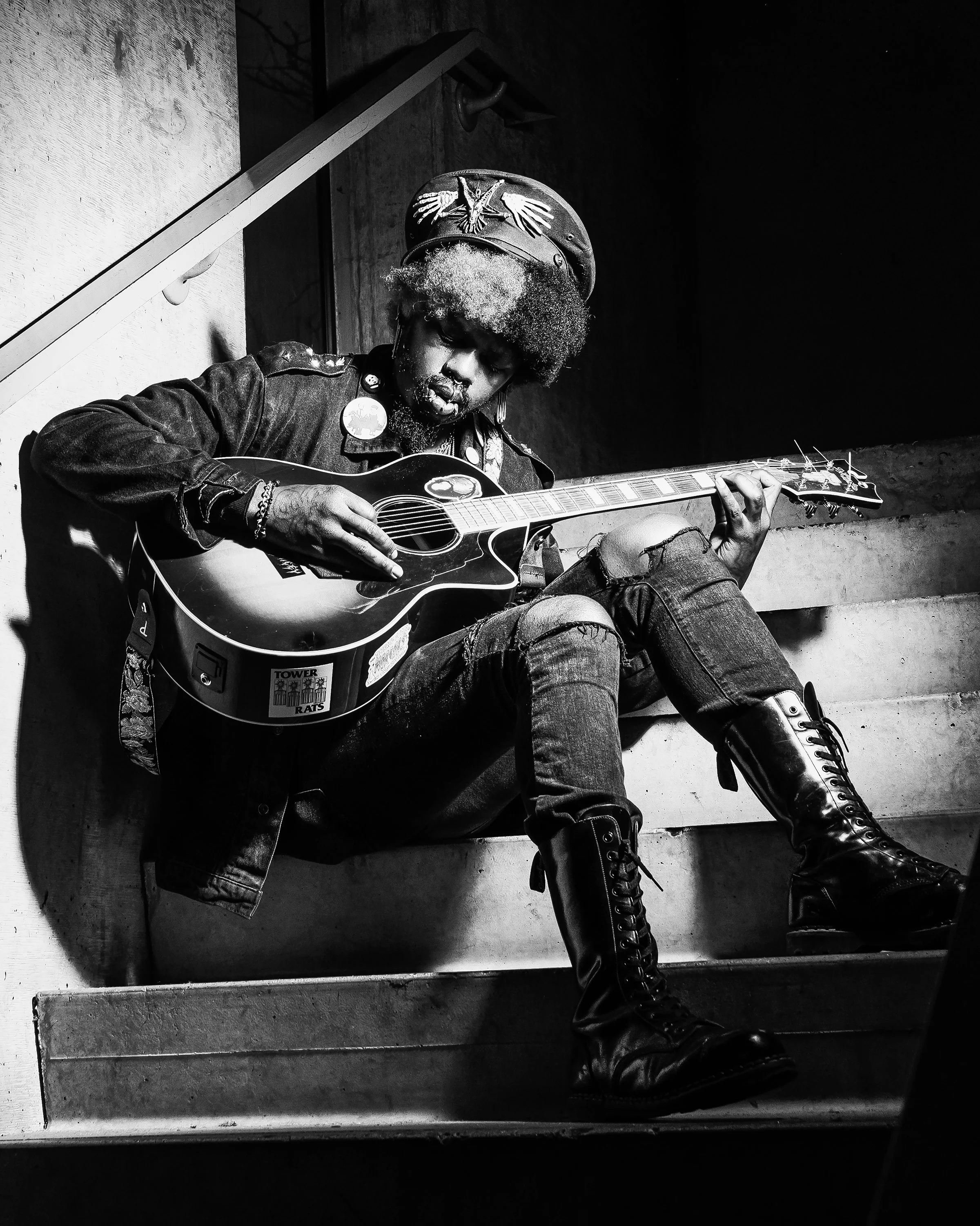 A person with curly hair, wearing a military-style hat, leather boots, and ripped jeans, playing an acoustic guitar while sitting on a step.
