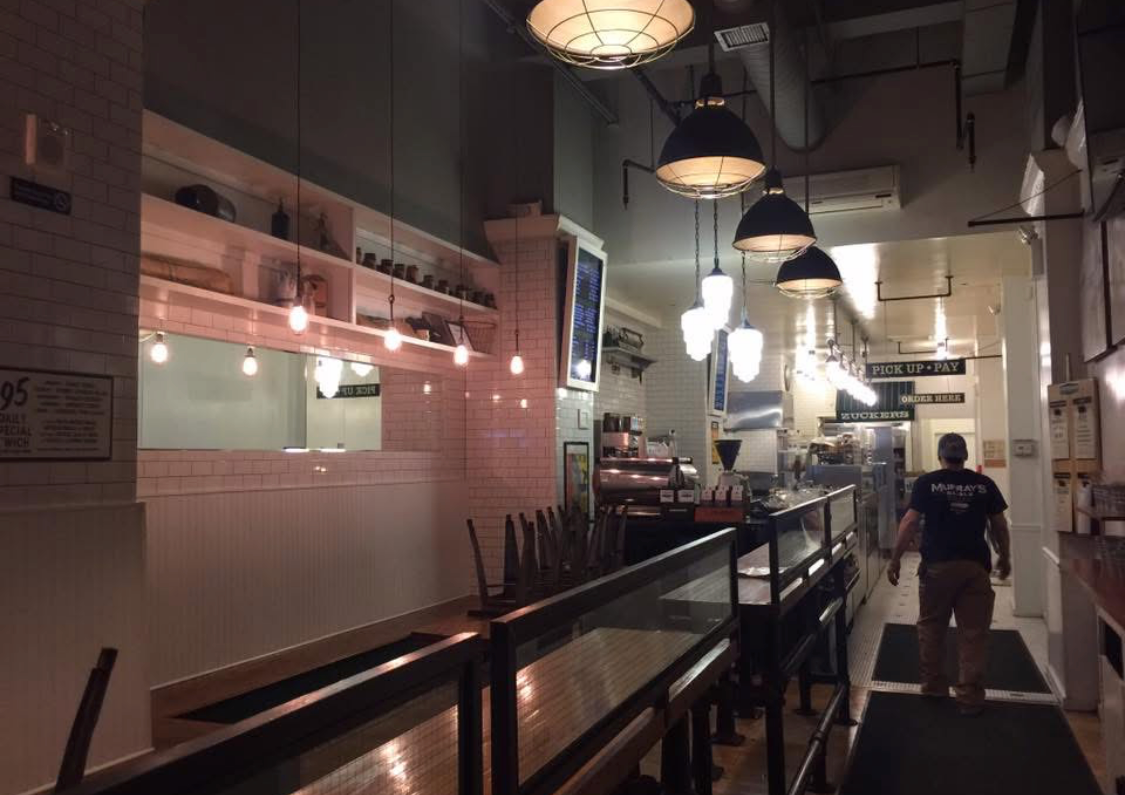 Interior of a modern restaurant or cafe with hanging pendant lights, a kitchen window, and a person walking toward the order pickup area.