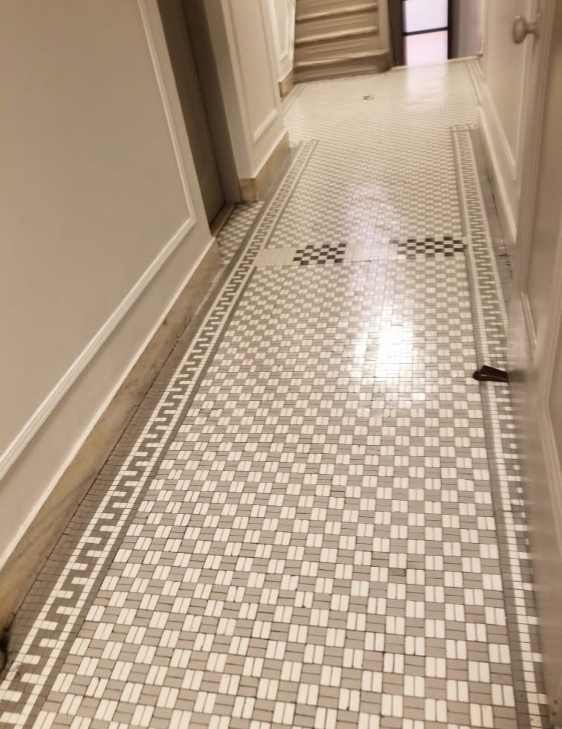 A narrow hallway with a mosaic tile floor featuring small white and beige tiles, with a decorative border and a black and beige checkered pattern near the center. There are stairs at the end of the hall and some debris on the floor.