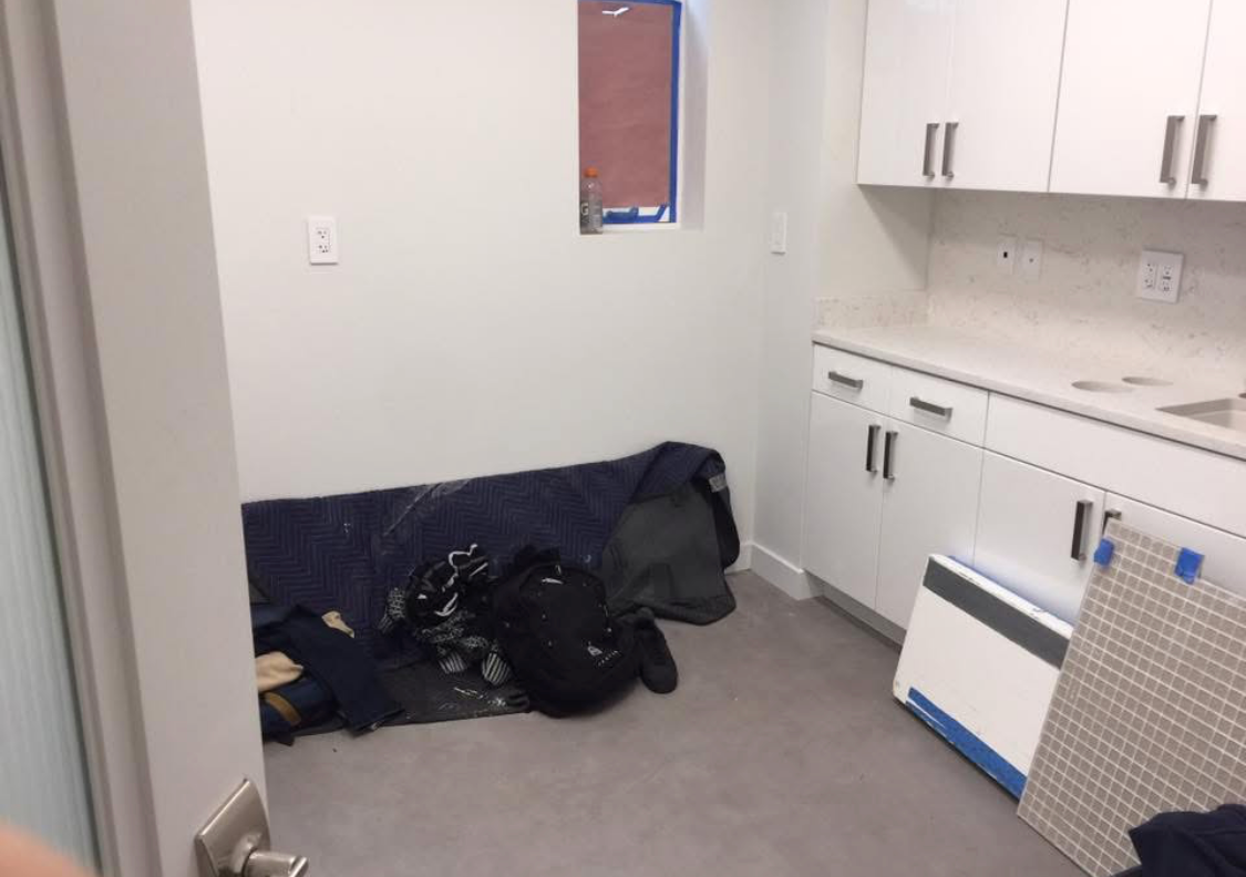 Room with a small window, white walls, white cabinets, and a gray floor. There are some bags and clothing on the floor, a white baseboard heater, and an image or panel leaning against the wall.