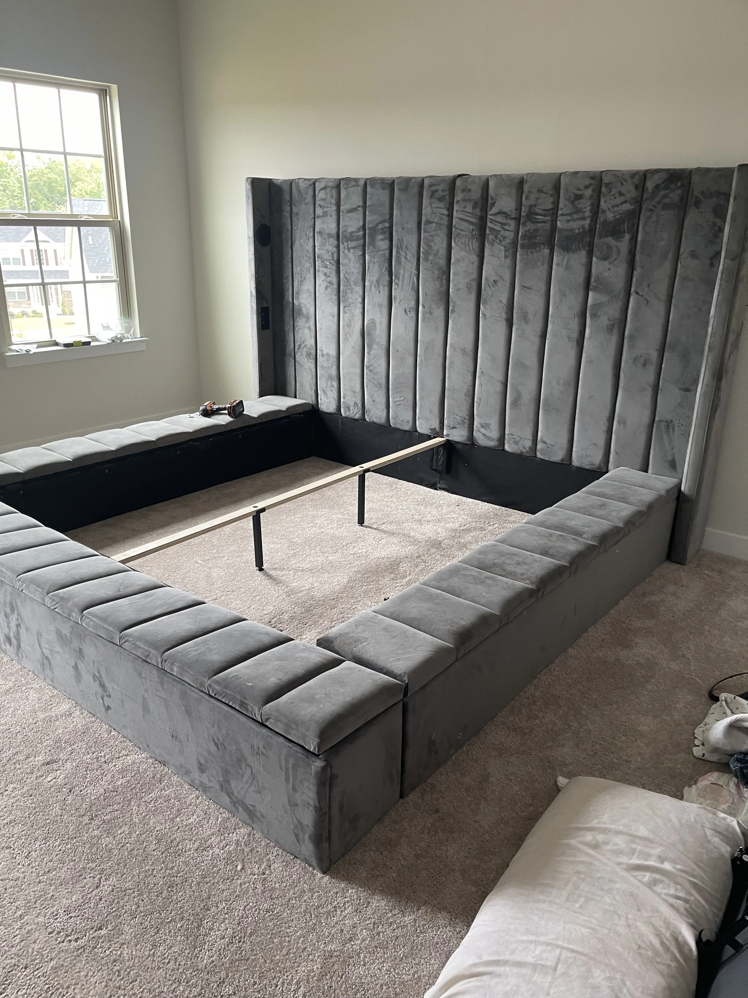 Unfinished room with a large, gray, velvet sofa frame arranged in a square shape on a beige carpet. A window on the left side allows natural light in. A small black and red drill is on the sofa frame.