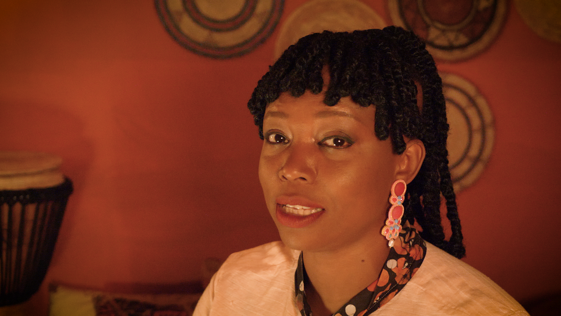 A woman with short twisted black hair, wearing pink statement earrings, a patterned shirt, and a cream-colored jacket, looks at the camera with a slight smile against a background of colorful circular wall art and a drum.