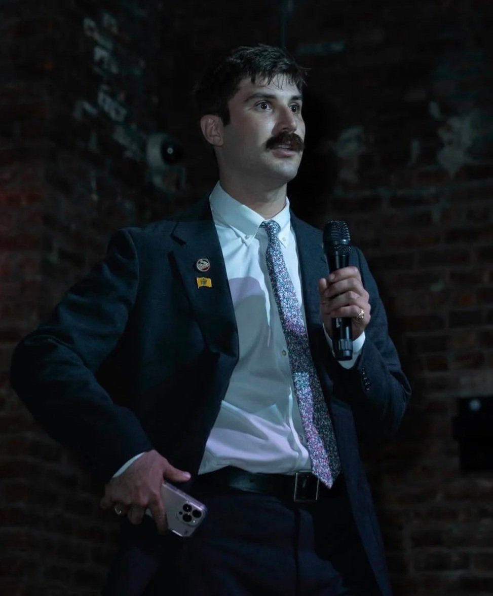 Ben Dziobek posing in front of a rocky barrier next to the ocean, wearing a jean jacket and white hoodie.