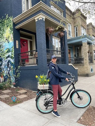 Person standing with a bike on a city sidewalk, wearing a cap and jacket, in front of a blue-painted building with a mural. The bike has a basket with yellow flowers.
