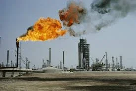 Oil refinery with burning flare stack and smoke against a clear sky.