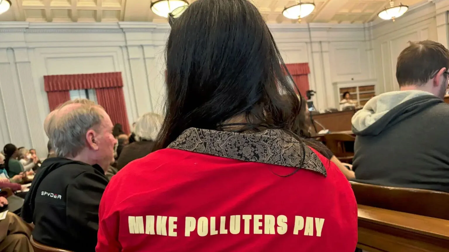Person wearing a red shirt with "Make Polluters Pay" text sitting in a courtroom.