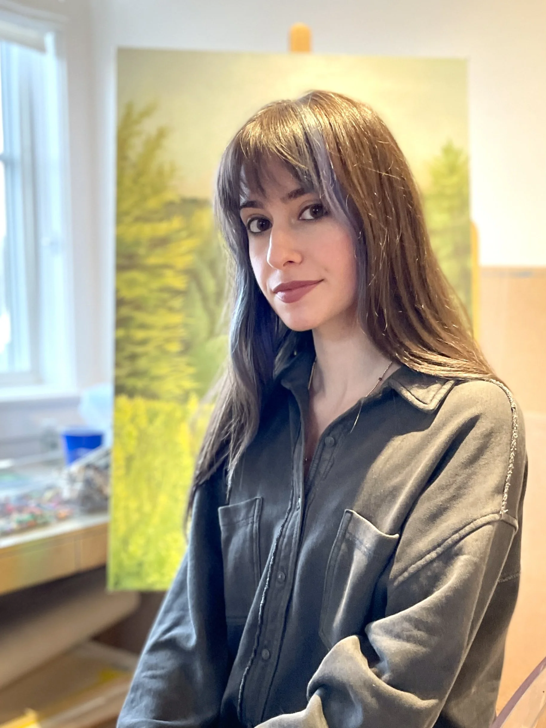 Person standing in front of a painting, wearing a button-up shirt, with long hair and bangs, in a room with natural light coming from a window.