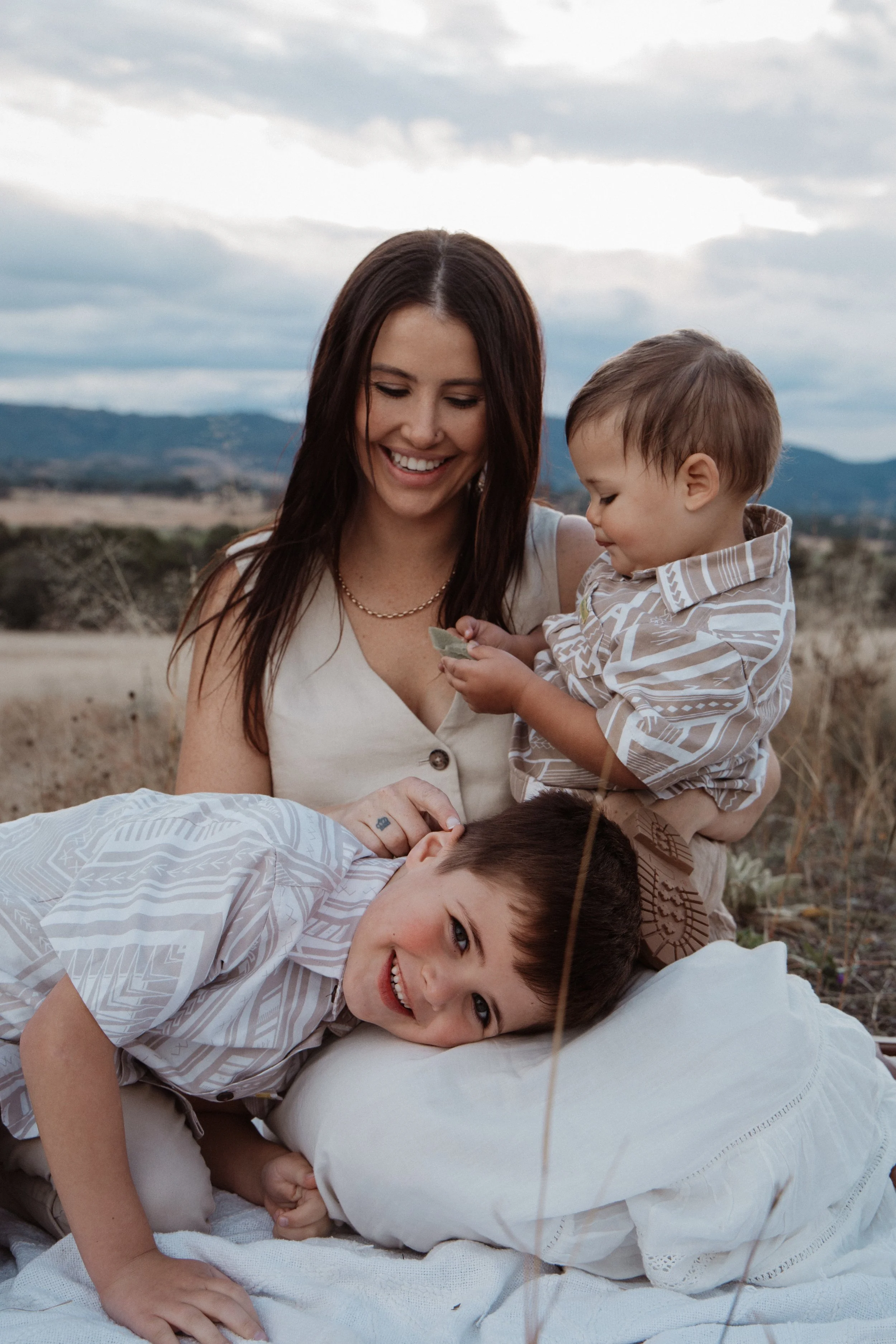 A smiling woman sits on a picnic blanket in a field with two young children. One child is sitting on her lap holding a leaf, while the other is lying next to her, smiling at the camera. The background features a landscape with hills and a cloudy sky.