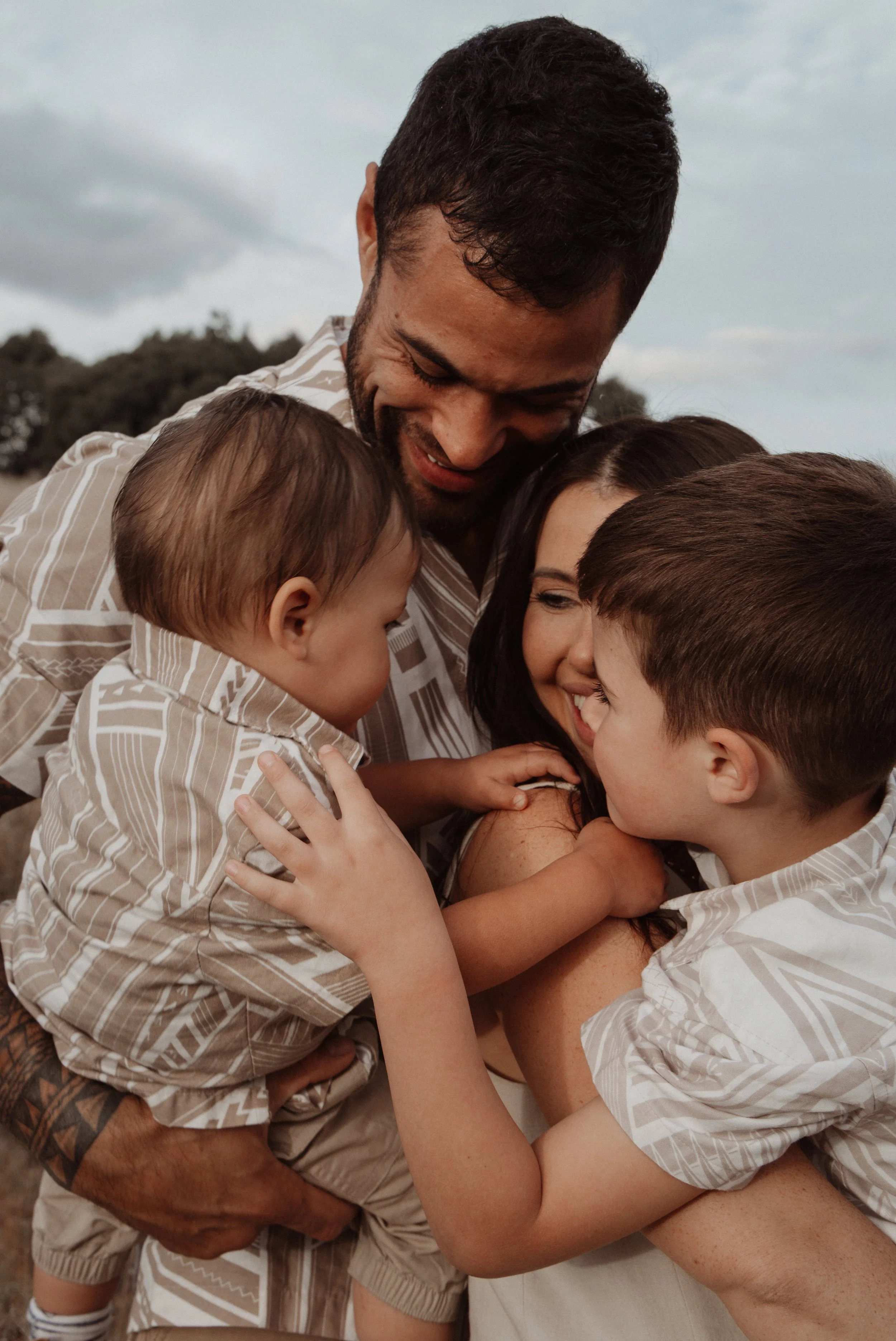A family of four embracing, dressed in matching patterned shirts. A man is holding a baby, while a woman smiles with an older child in front of her. Outdoor setting.