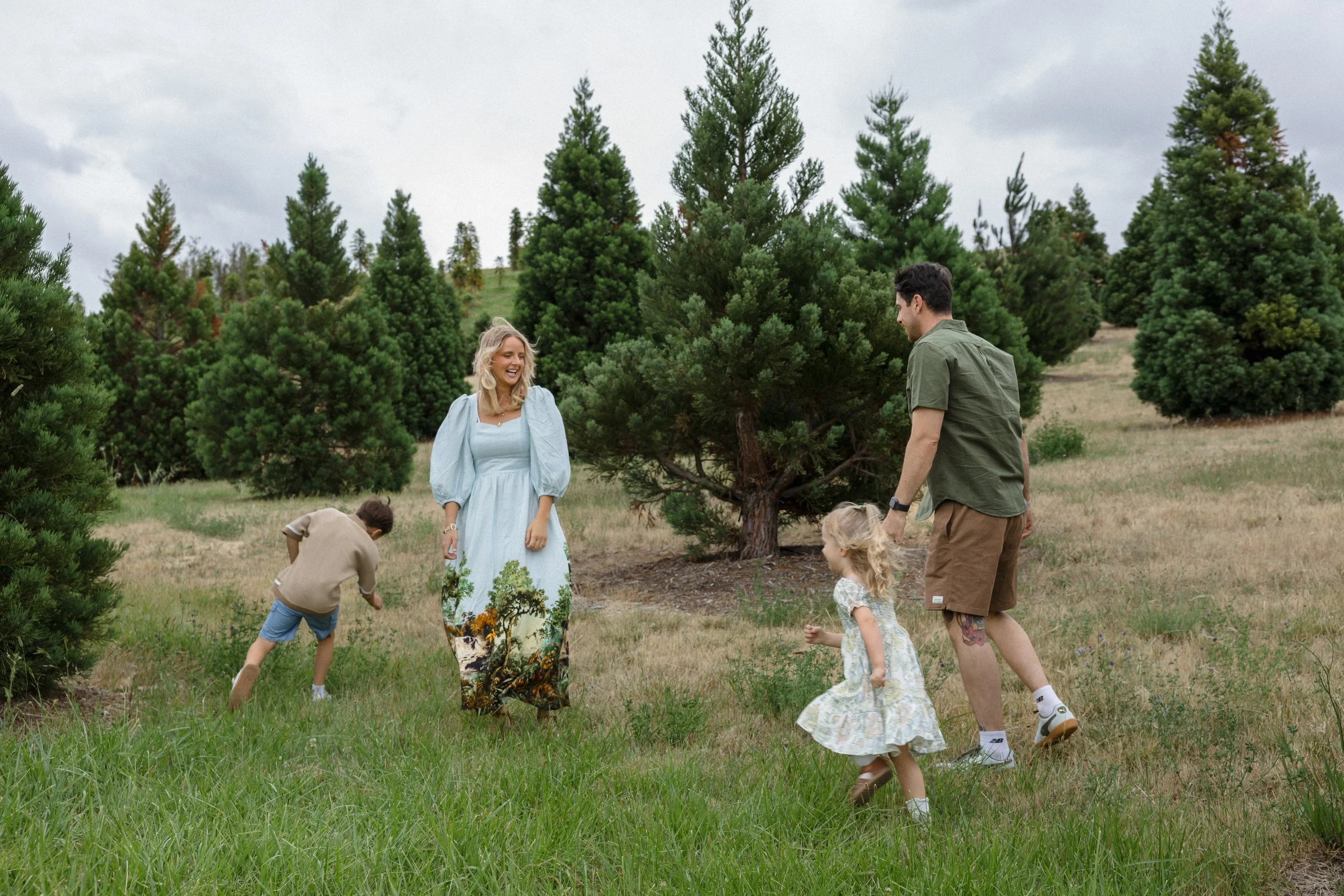 A family of five enjoying a day outdoors among pine trees on a cloudy day. The mother wears a long, light blue dress with a scenic nature print at the bottom and is smiling. The father wears a green short-sleeve shirt and brown shorts, holding hands with a young blonde girl in a floral dress. A young boy in shorts and a brown sweater and another girl in a light-colored dress are also playing nearby.