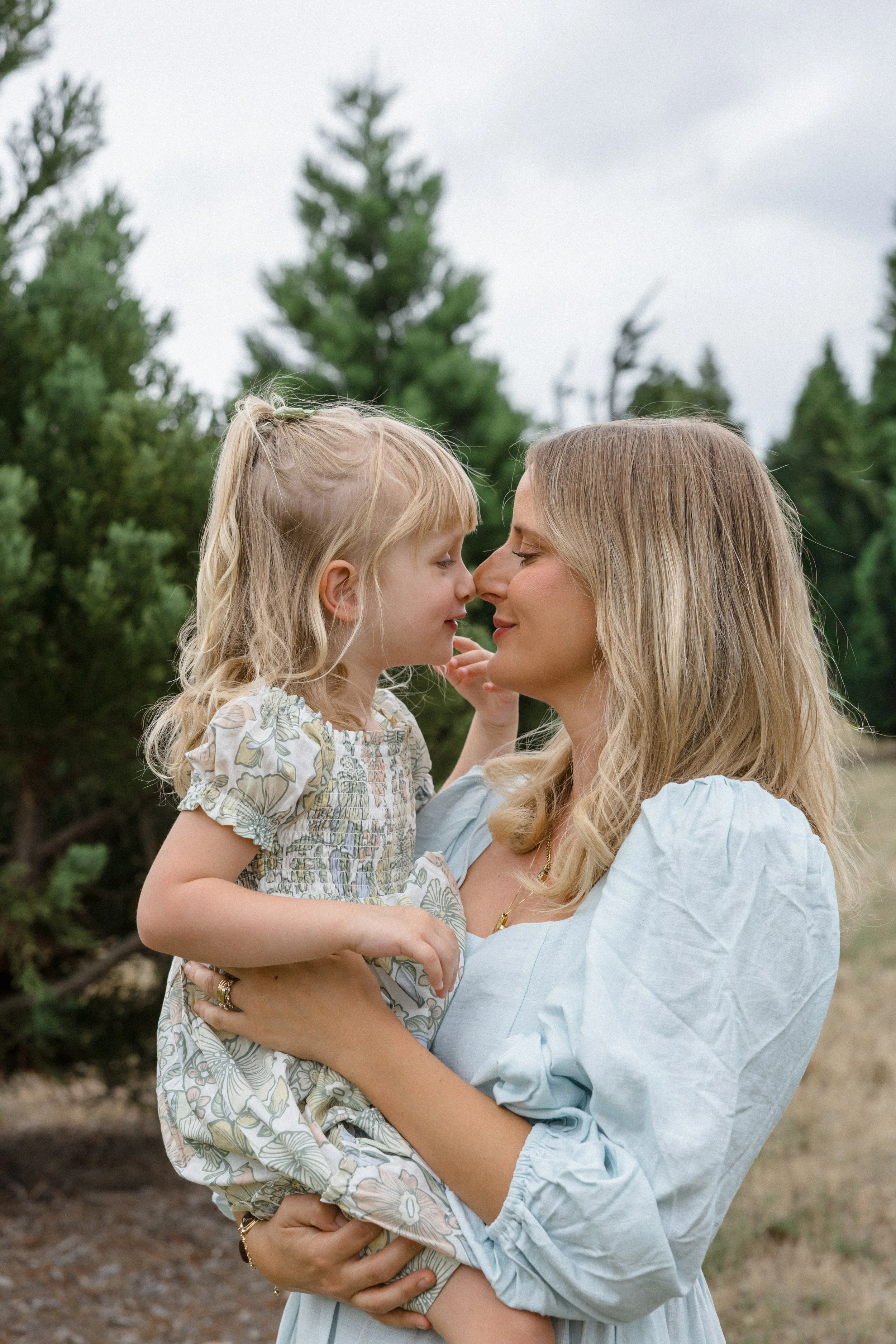 A woman holding a young girl outdoors, with trees in the background, their faces close together.