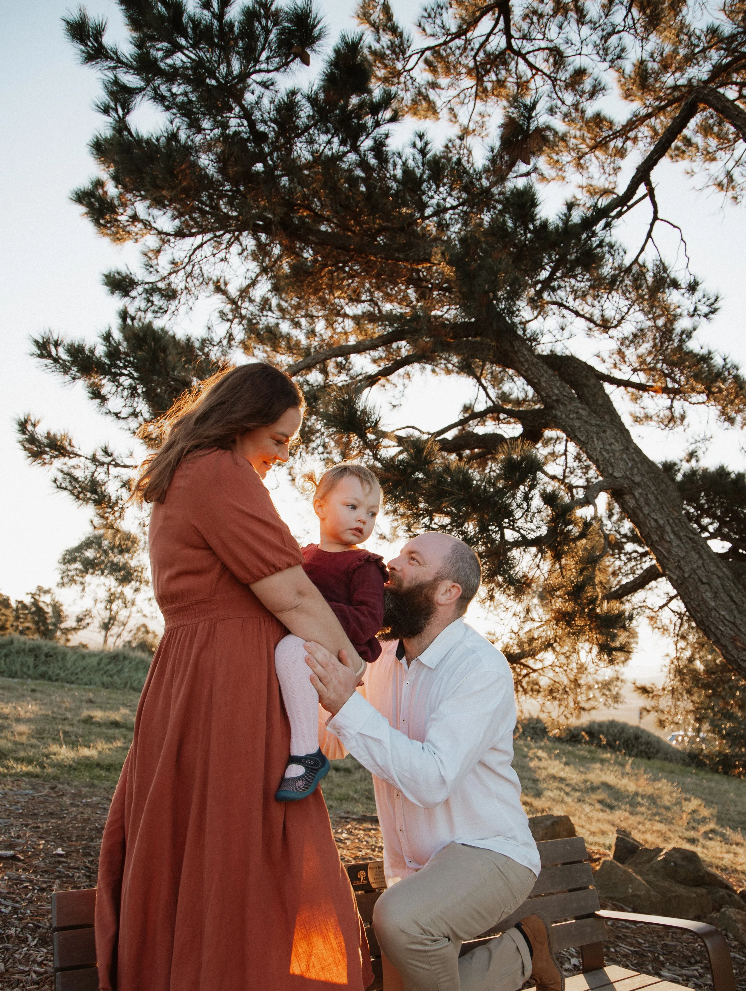 A family outdoors with a woman in a long dress holding a child while a man kneels beside them, under a large tree during sunset.