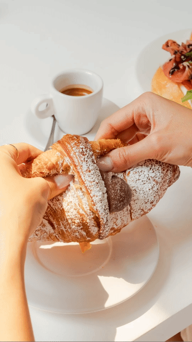 Hands pulling apart a powdered sugar croissant on a white plate, with a cup of espresso and food in the background.