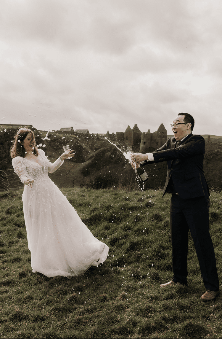 Bride and groom celebrating their elopement in Ireland outdoors with champagne on a grassy hill with castle ruins in the background, as champagne sprays between them