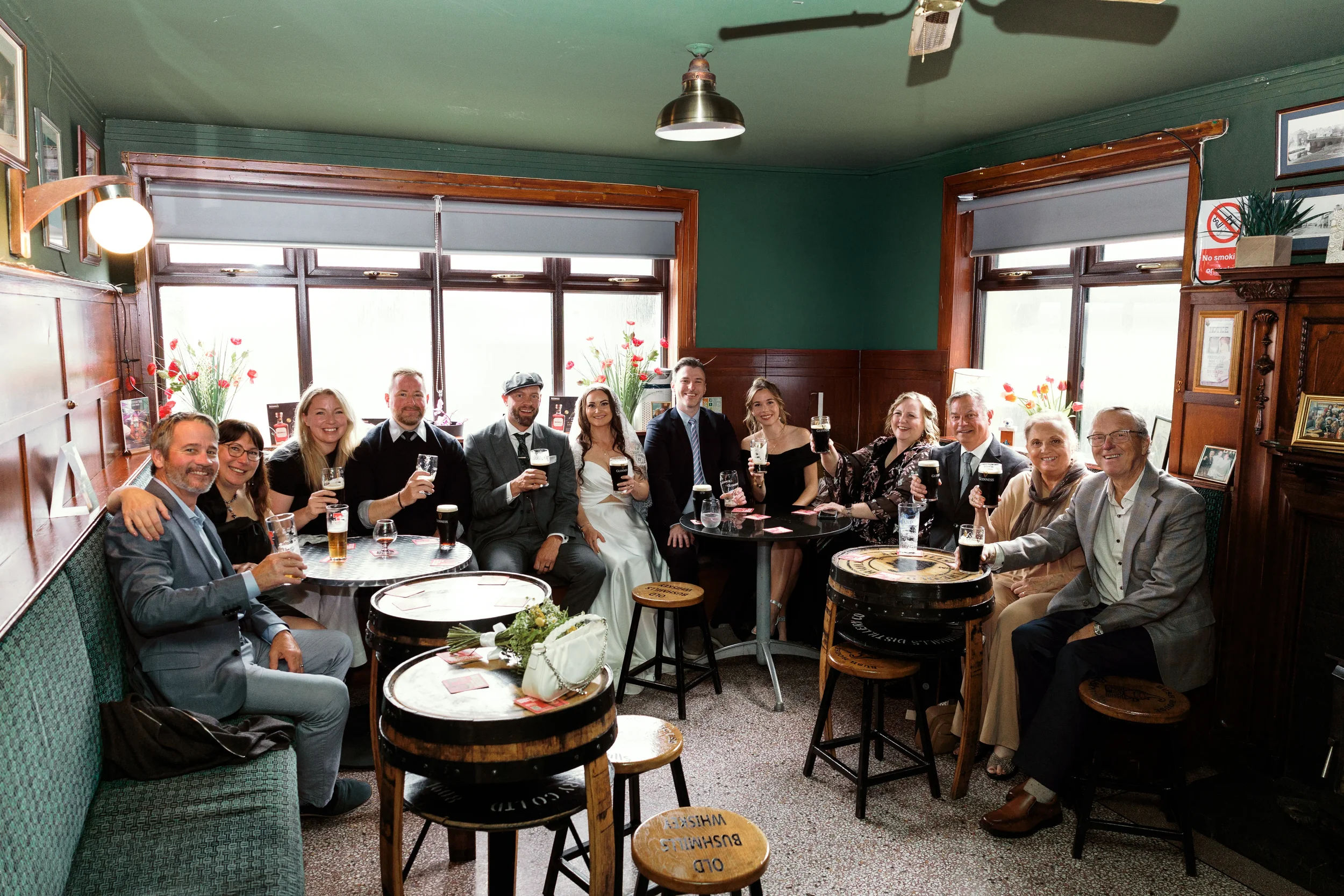 Group of people celebrating, holding drinks, sitting at tables in a cozy pub or restaurant with green walls, large windows, and wooden furniture.