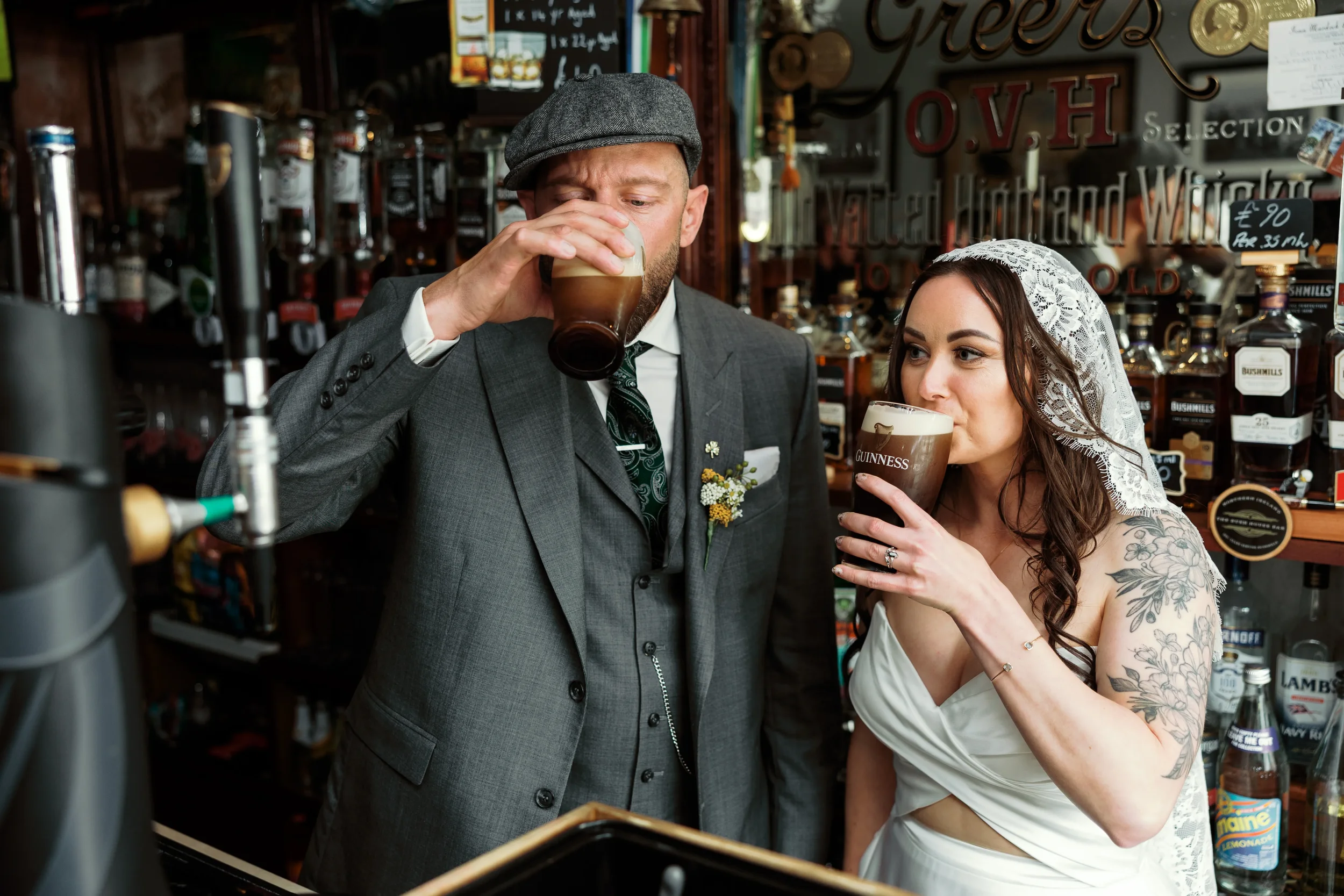 A man in a grey suit and cap and a woman in a wedding dress with tattoos are drinking pints of Guinness beer in a bar.