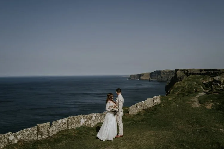 A couple in wedding attire standing on a grassy area near a stone wall, overlooking the ocean and cliffs in the distance during their destination wedding in Ireland