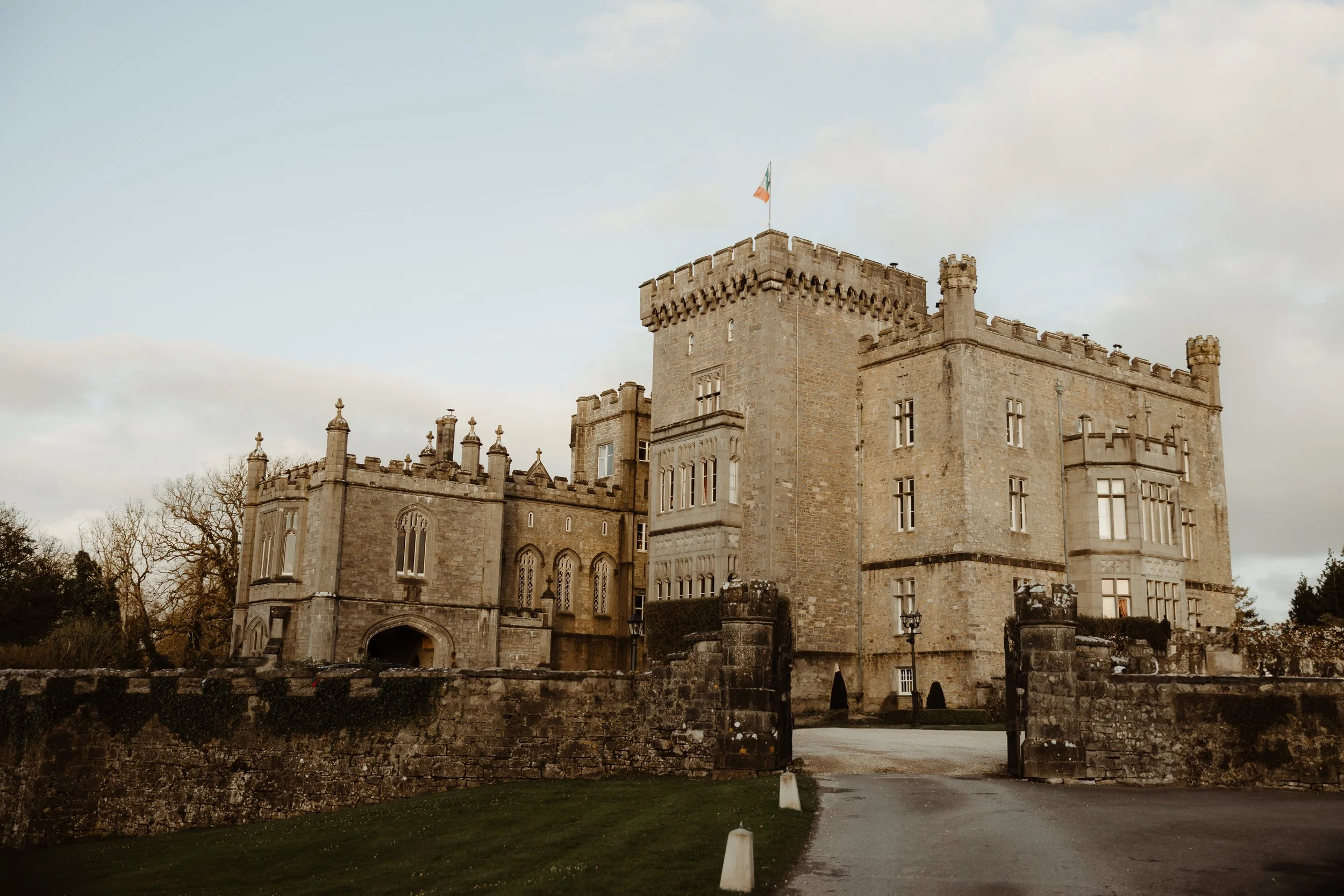 An expansive view of the Gothic stone facade of Markree Castle in Ireland under an overcast sky, featuring crenellated battlements, a central tower with an Irish flag, and a stone-walled driveway in the foreground.