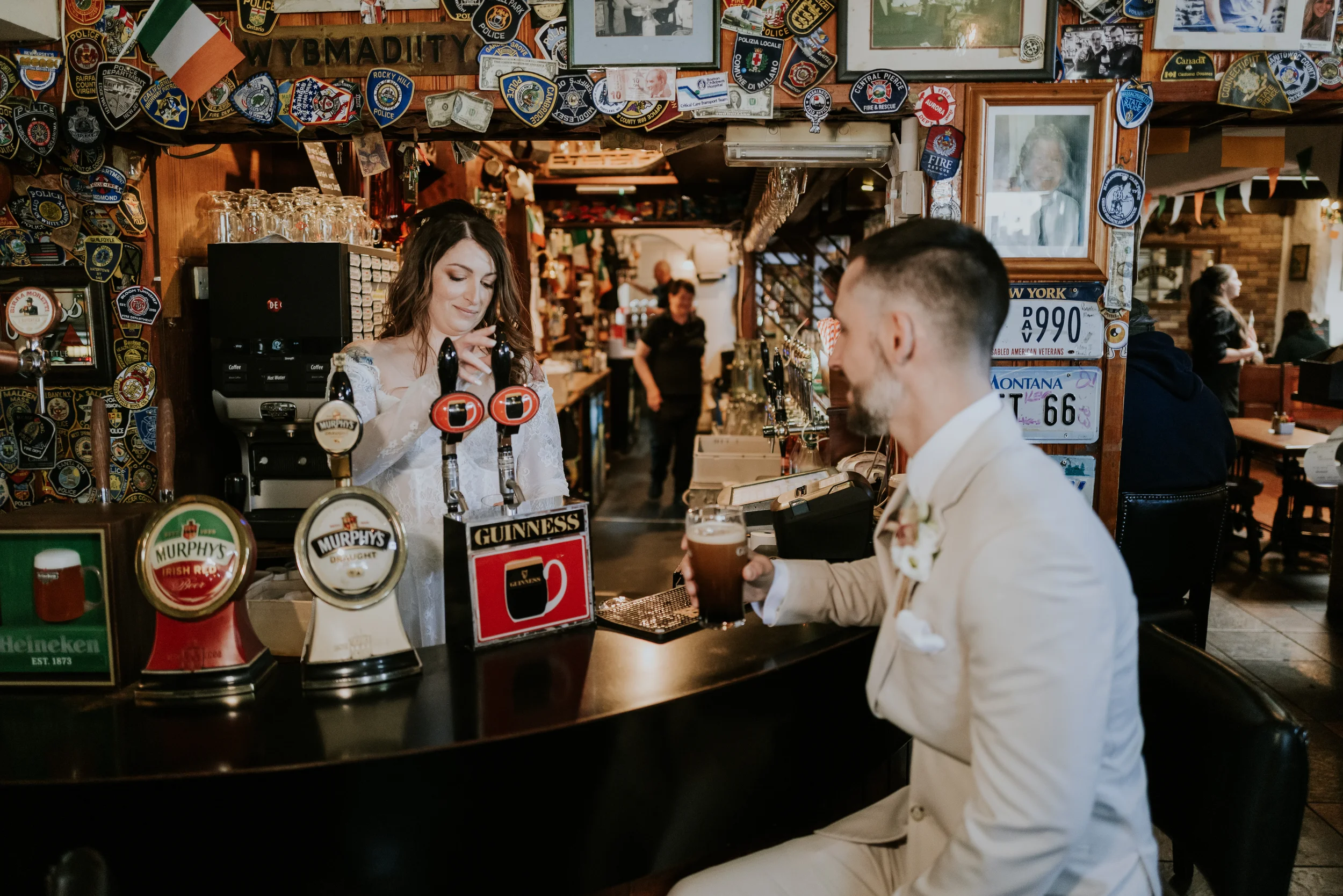 A man in a white suit with a boutonniere sitting at a bar, holding a pint of beer, while a woman behind the bar pours a drink. The bar is decorated with various patches and signs, and other patrons are visible in the background.