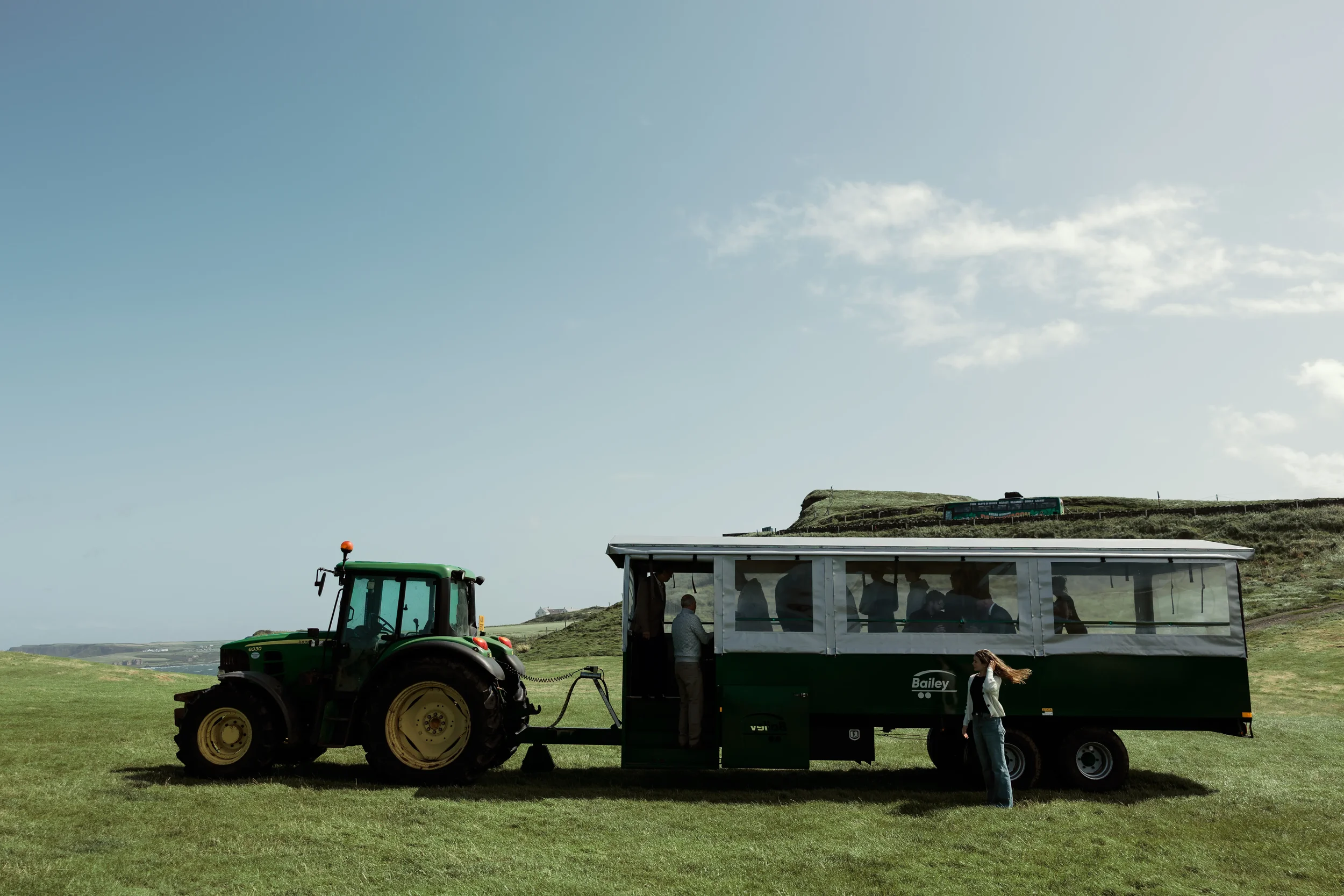 A large green tractor attached to a mobile lounge trailer with people inside, parked on a grassy field with a hilly landscape and a train in the background under a partly cloudy sky.