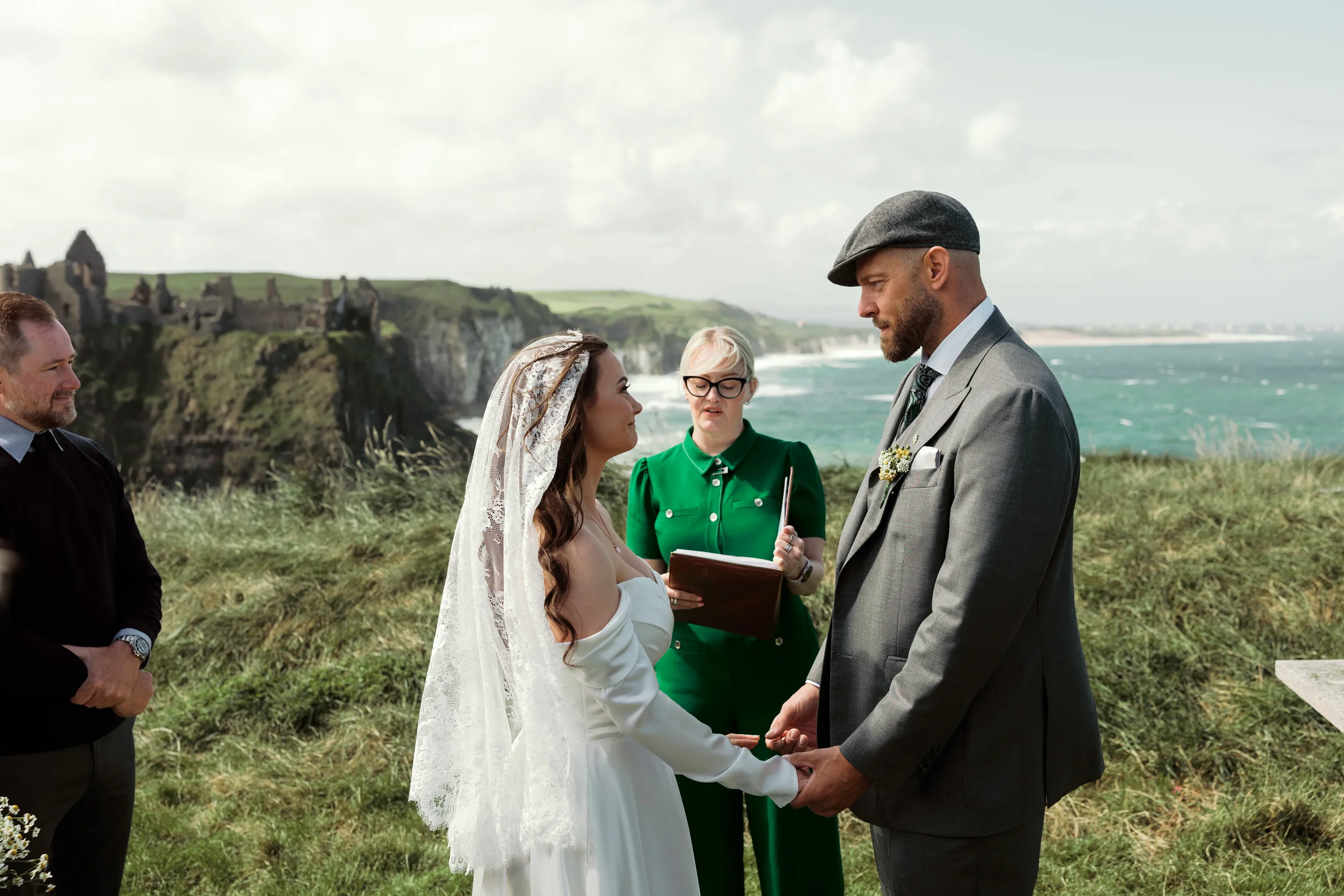 A bride and groom holding hands during an outdoor wedding ceremony by the coast, with an officiant standing between them and an onlooker on the left, cliffs and ocean in the background.