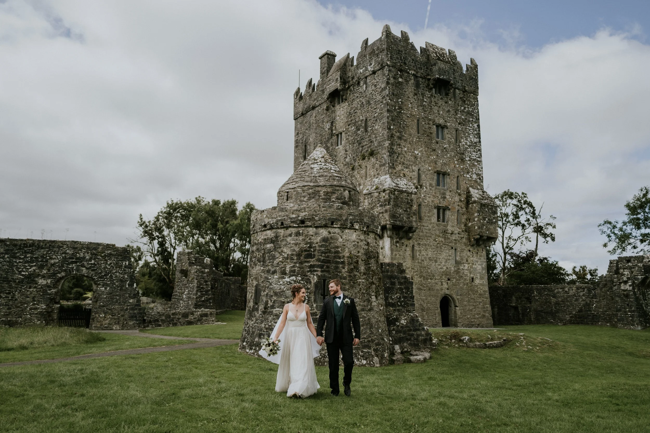 Bride and groom in wedding attire with Scottish kilts, standing on a grassy hill overlooking a coastline with cliffs and ruins, under a partly cloudy blue sky.