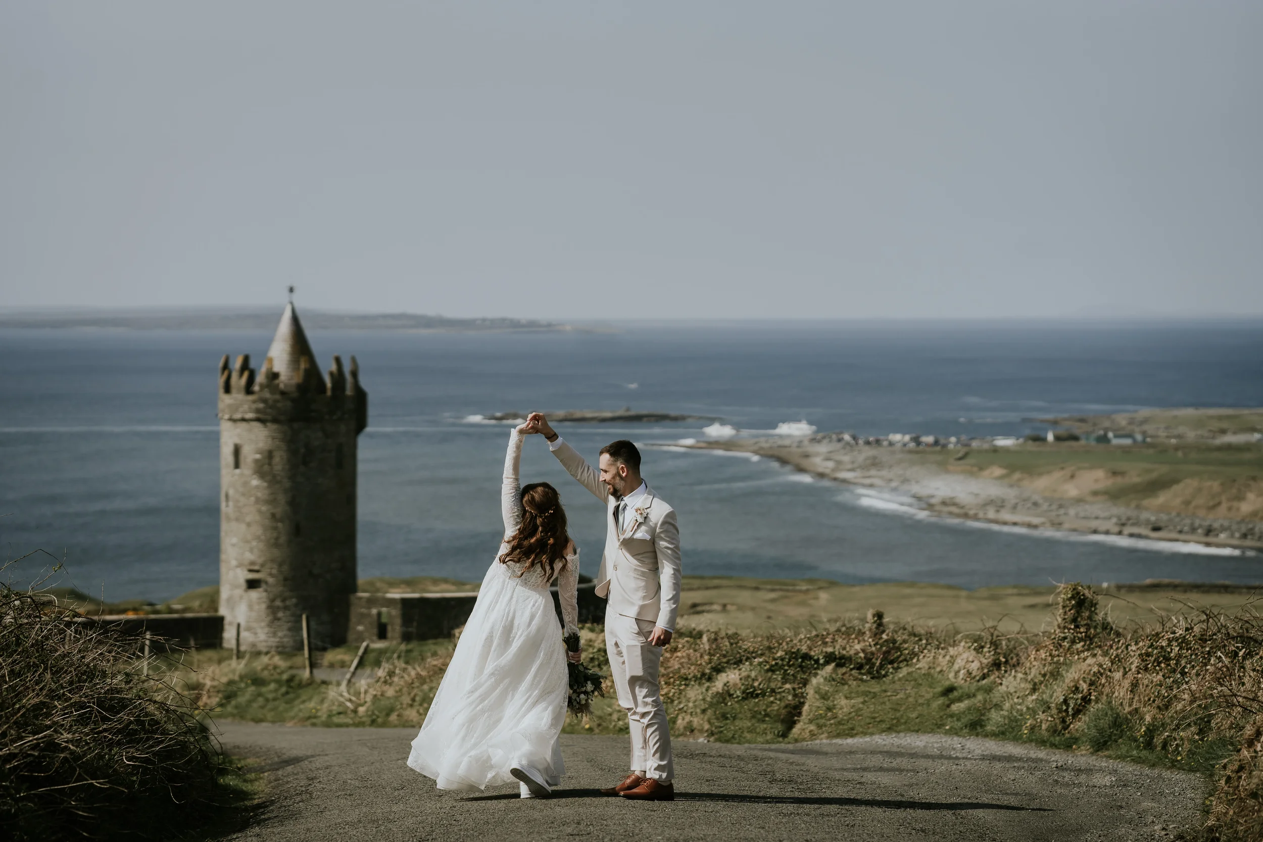 A bride and groom dancing on a rural road with a castle and ocean in the background.