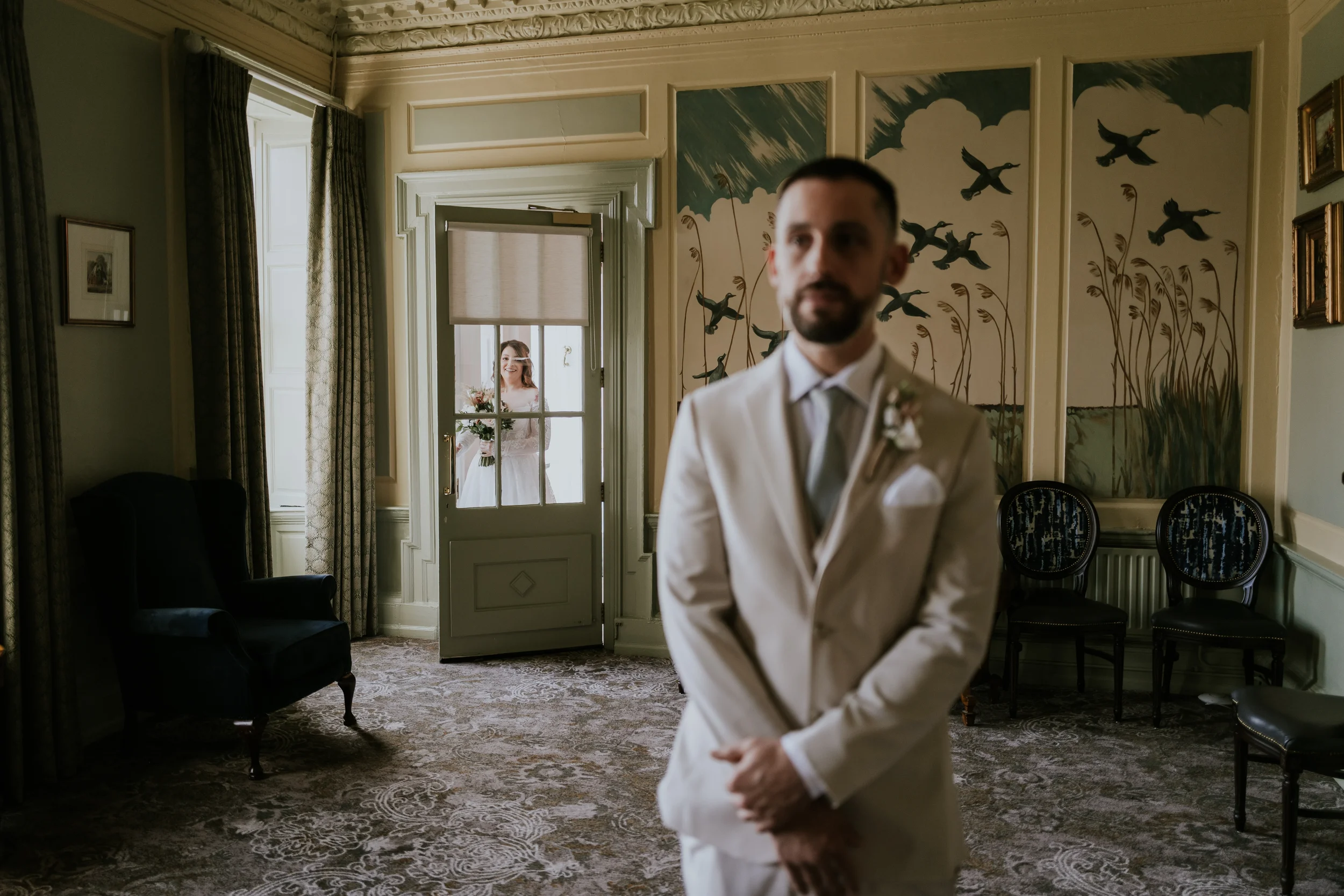 A groom in a white suit with a boutonniere stands in a decorated room, while a bride in a wedding dress holding a bouquet looks through a window at him.